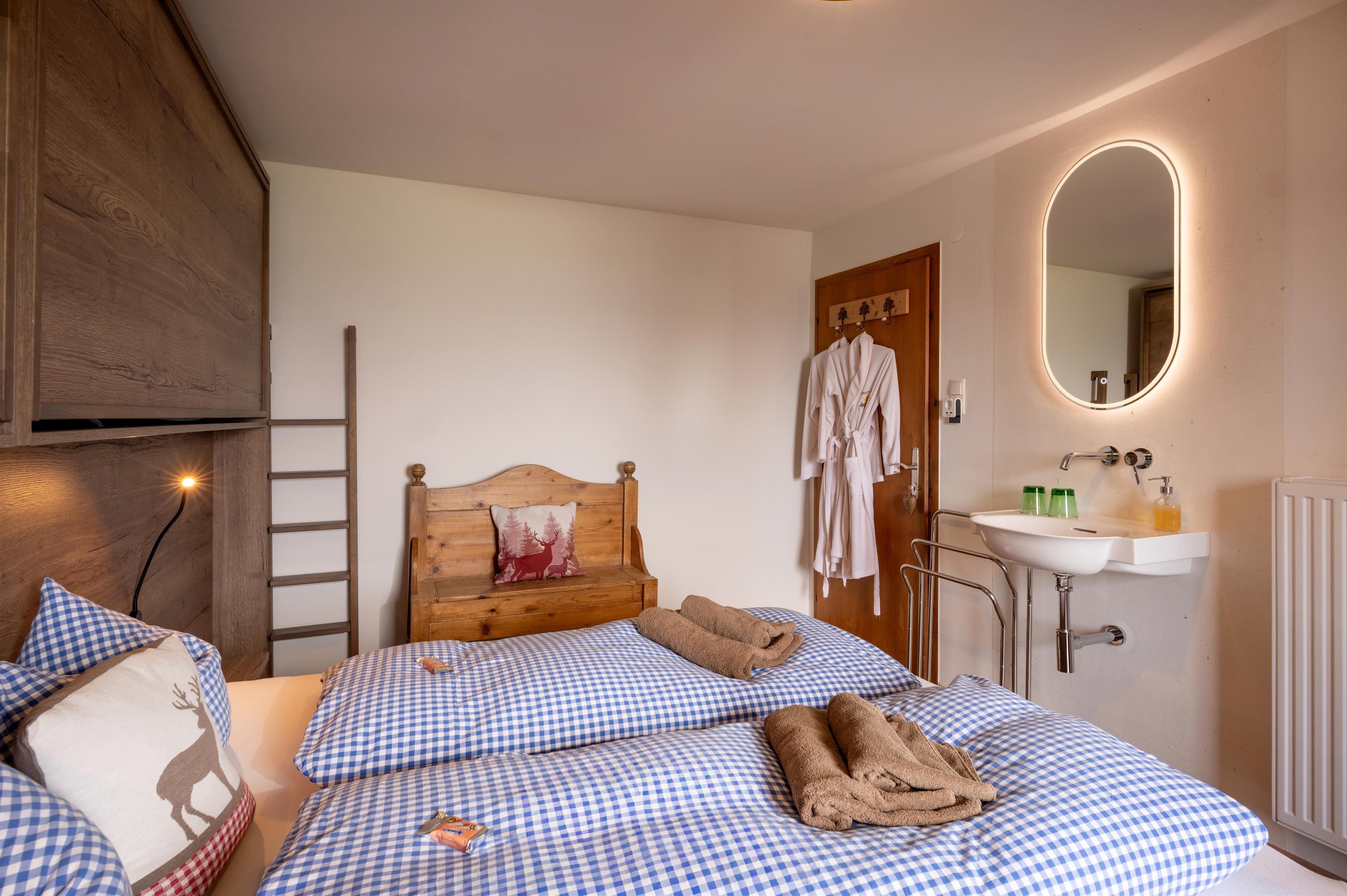 Ein gemütliches Schlafzimmer mit einem Doppelbett und blau-weißen Karos. Es gibt einen Sessel, ein Waschbecken und einen großen Spiegel an der Wand.