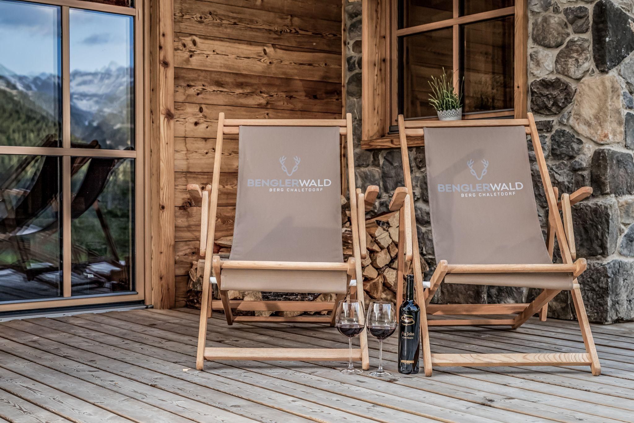 Zwei entspannende Liegestühle stehen auf einer Holzterrasse vor einen Chalet. Daneben stehen zwei Weingläser und eine Flasche Wein.