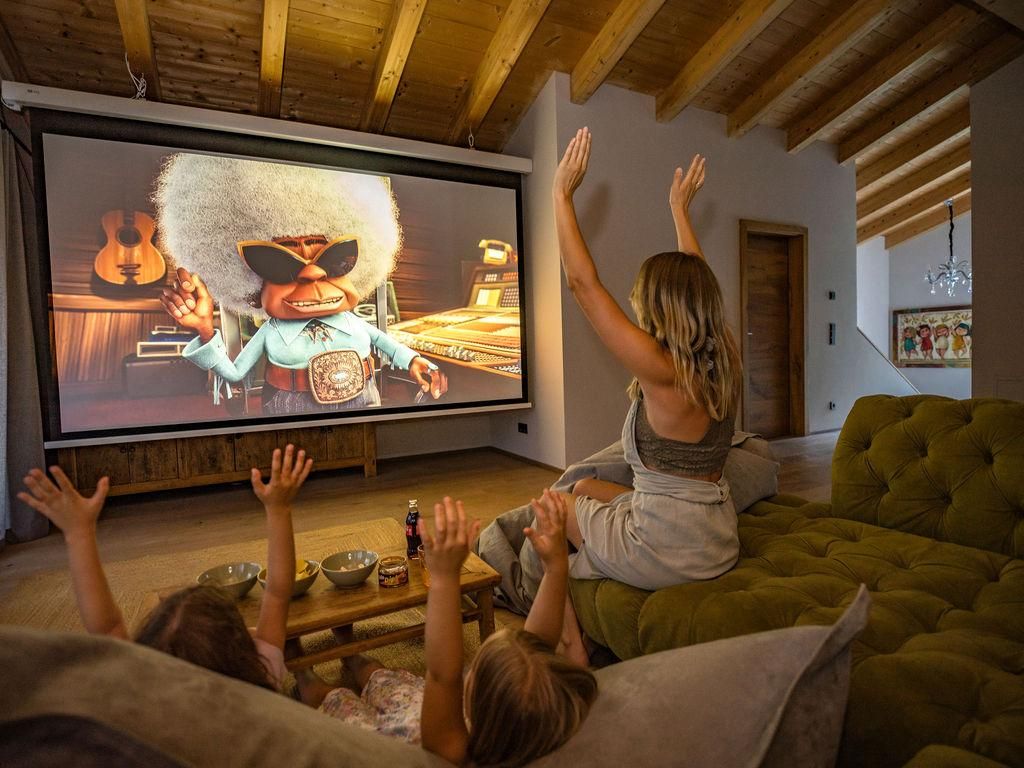 Eine Familie schaut begeistert einen Animationsfilm auf einer großen Leinwand. Die Erwachsenen und Kinder heben ihre Hände, um mit dem Charakter im Film zu interagieren.