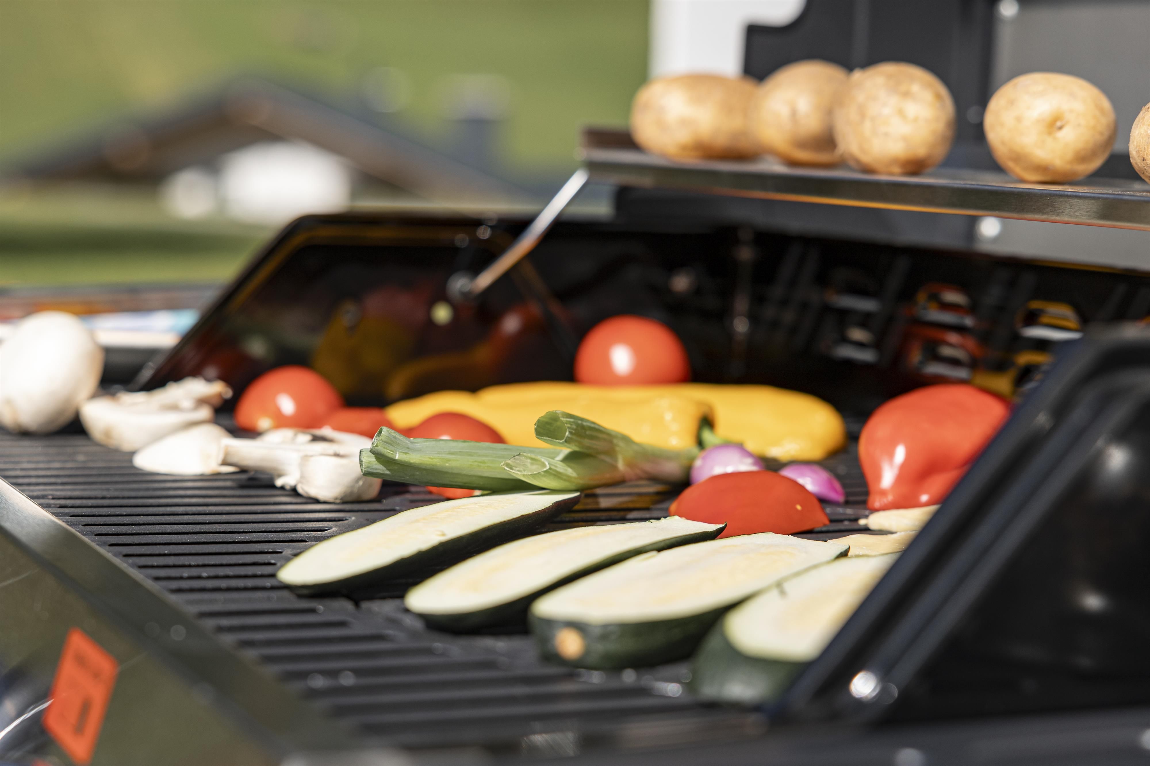 Buntes Gemüse auf einem Grill mit frischen Zutaten wie Zucchini, Tomaten und Paprika. Im Hintergrund sind Kartoffeln sichtbar, die für das Grillen vorbereitet werden.