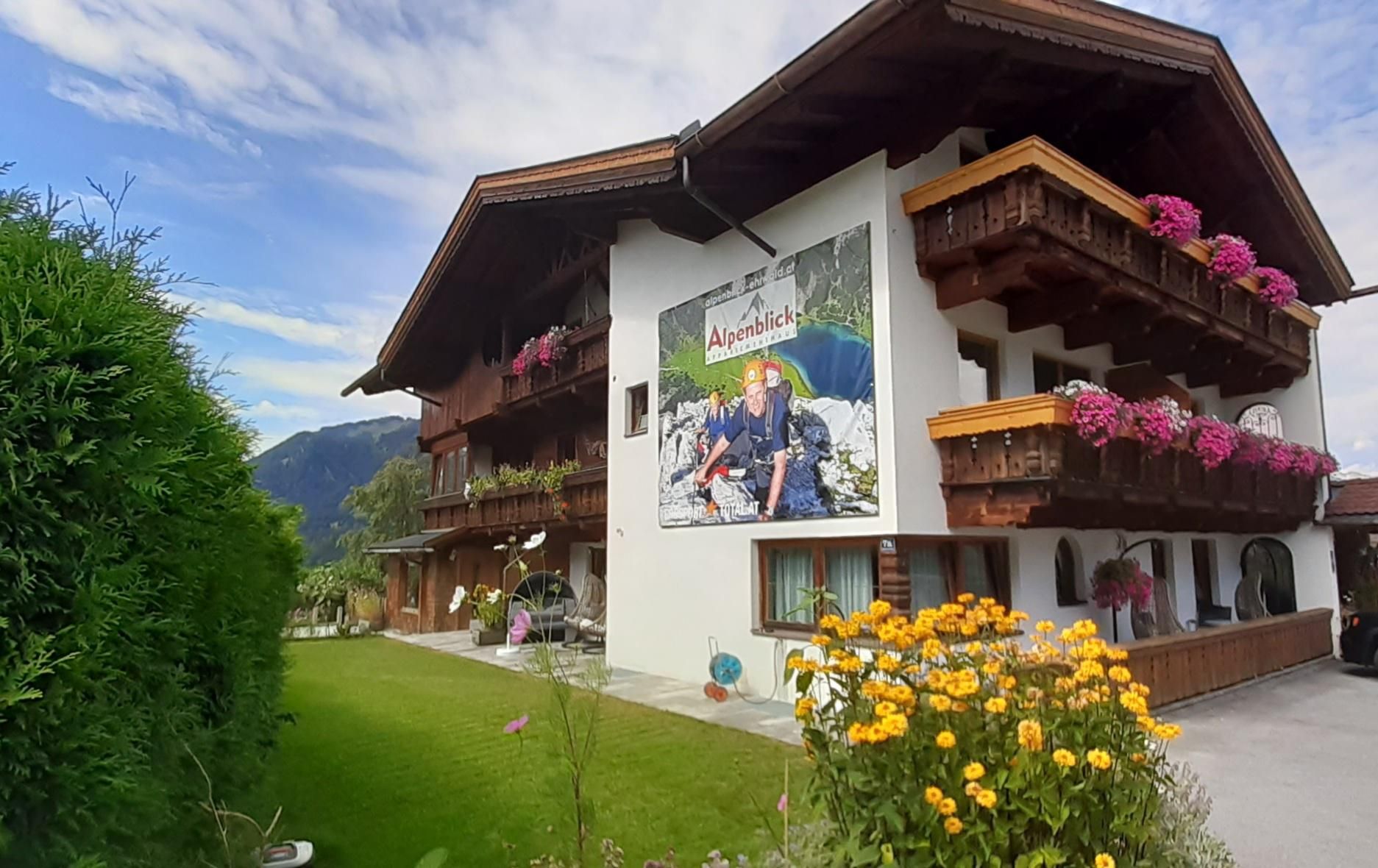 Ein malerisches Gebäude im Alpenstil mit blühenden Blumen und einem einladenden Garten. An der Fassade hängt ein großes Plakat, das Gäste anlockt.