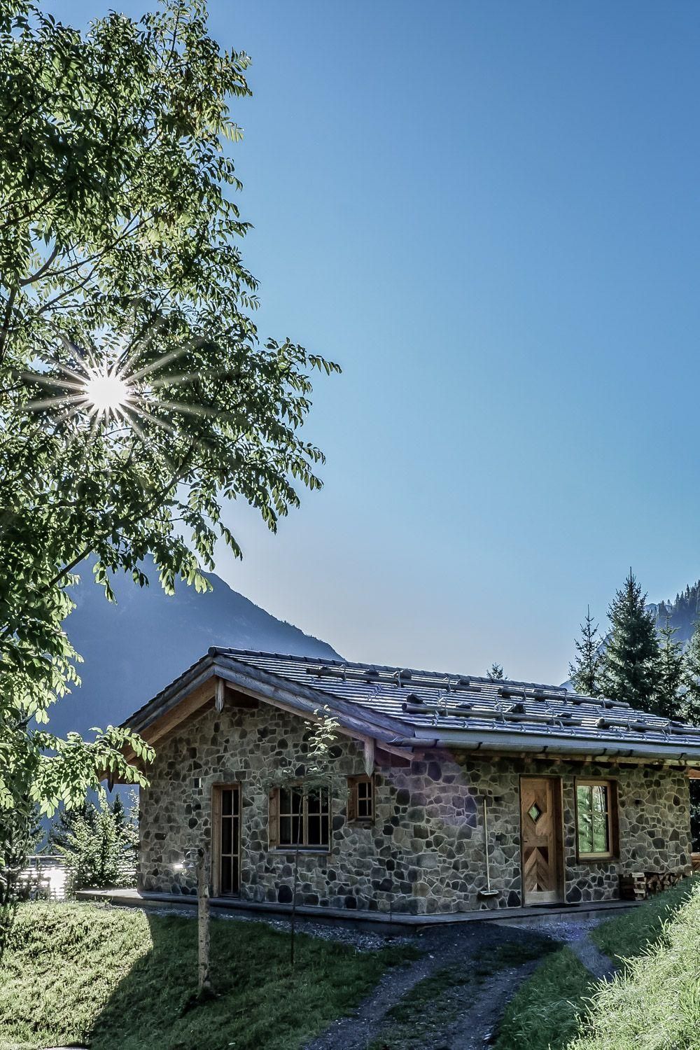 Stein Chalet umgeben von Bäumen und grüner Wiese. Die Sonne scheint auf das Dach und die umliegende Landschaft.