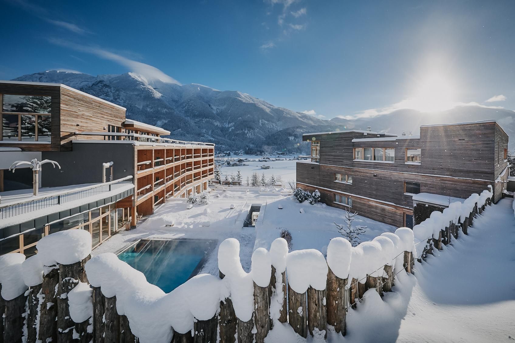Eine verschneite Winterlandschaft mit modernen Holzgebäuden und einem Pool. Im Hintergrund sind Berge und ein klarer Himmel zu sehen.