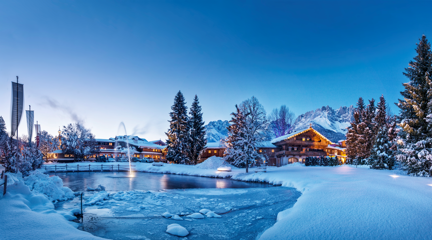 Eine malerische winterliche Landschaft mit schneebedeckten Bäumen und einem gefrorenen Teich. Im Hintergrund sind gemütliche Hütten und Berge unter einem klaren, blauen Himmel zu sehen.