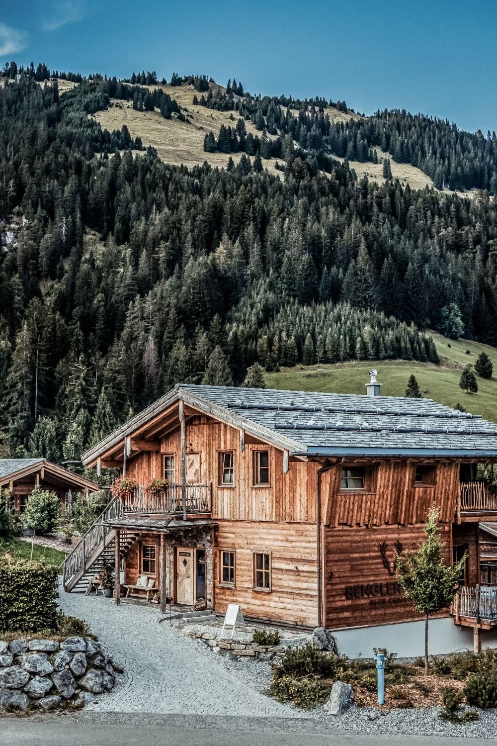 Ein gemütliches Holzhaus umgeben von grünen Wäldern und Bergen. Der Himmel ist klar und sonnig.