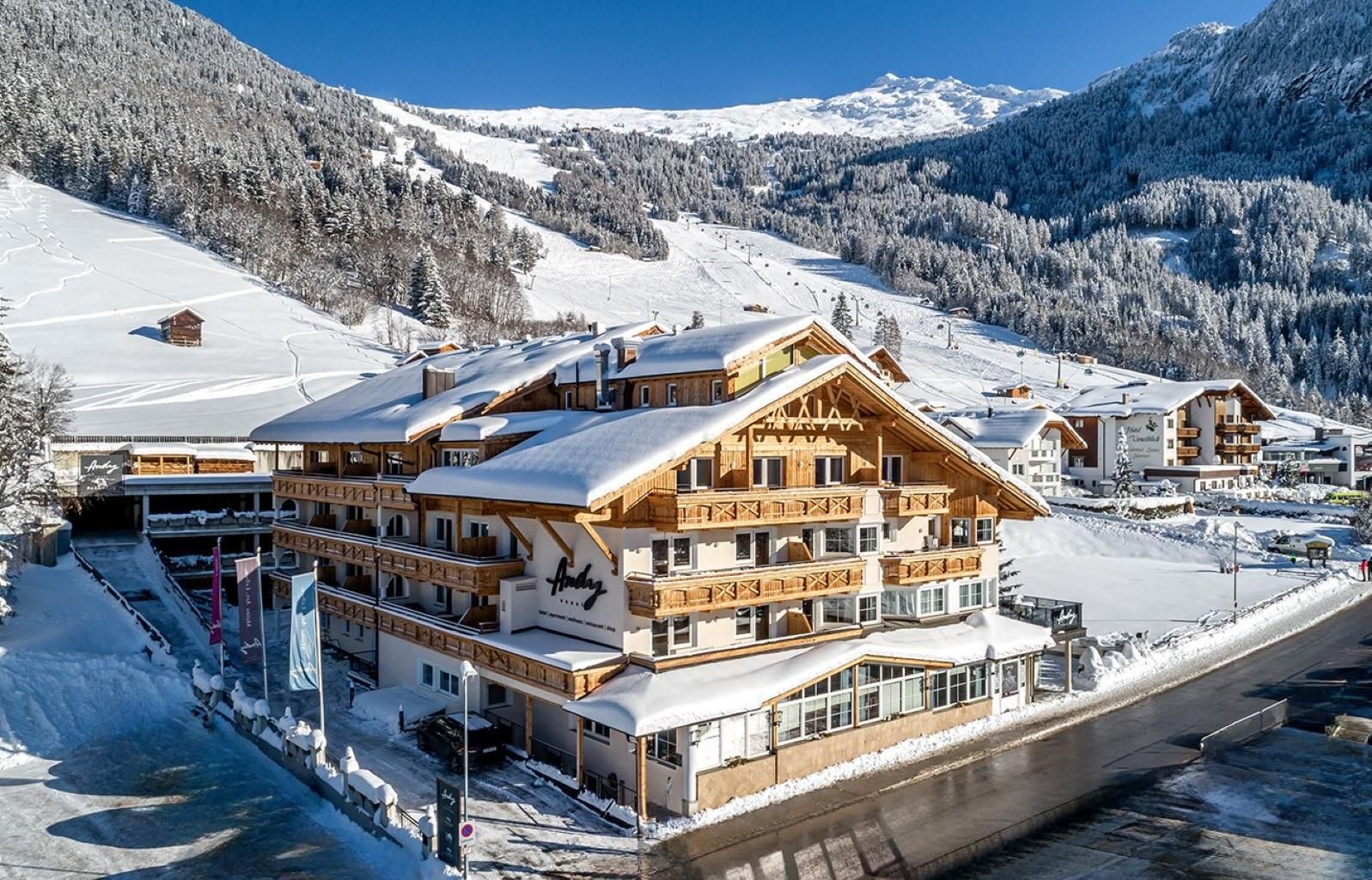 Ein charmantes Hotel im Schnee, umgeben von Bergen und Skigebieten. Der klare blaue Himmel verleiht der winterlichen Landschaft eine malerische Atmosphäre.