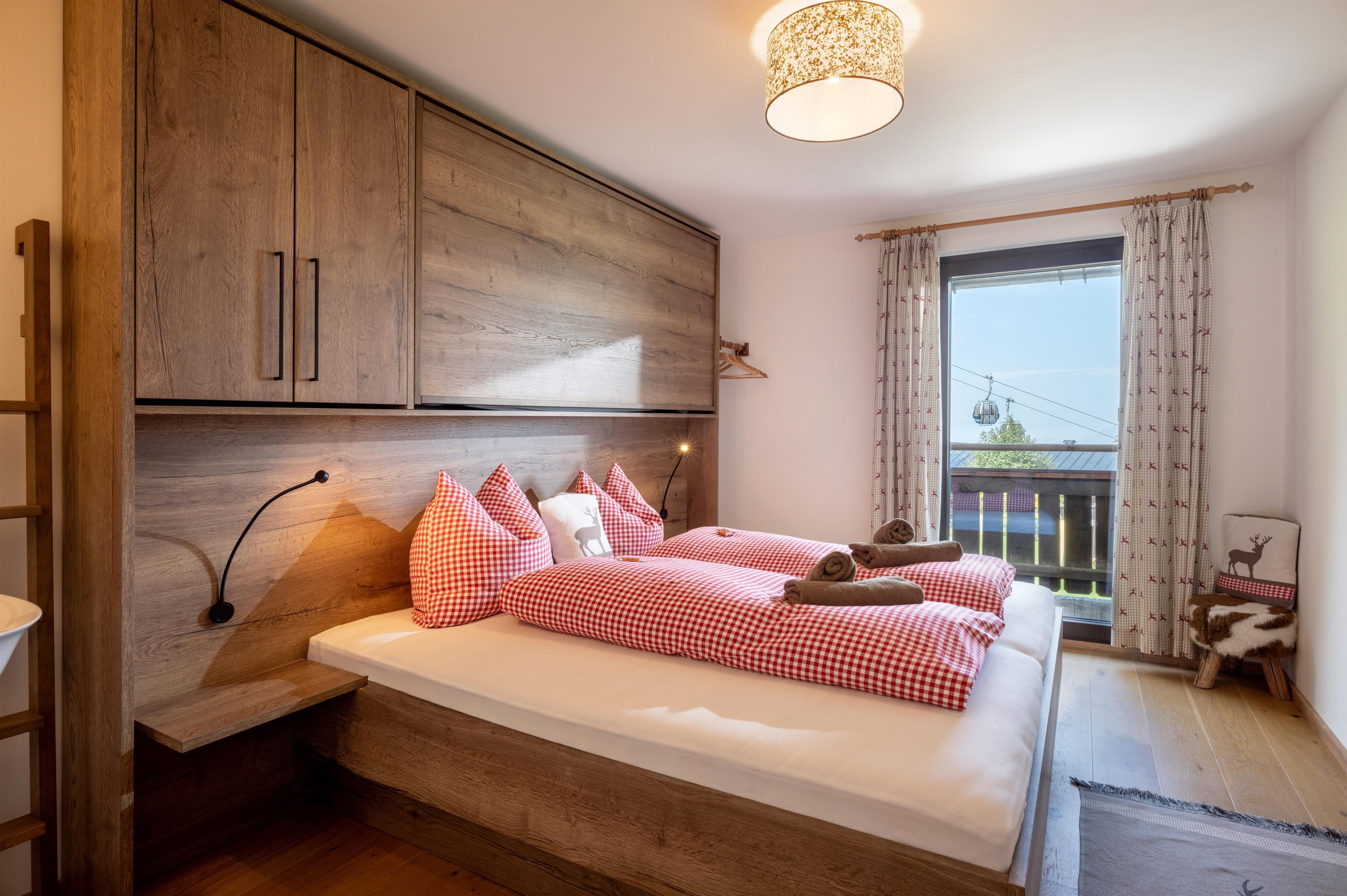 Ein gemütliches Schlafzimmer mit einem Doppelbett und rot-weißen Kissen. Das Zimmer hat große Fenster und moderne Holzmöbel, die eine einladende Atmosphäre schaffen.