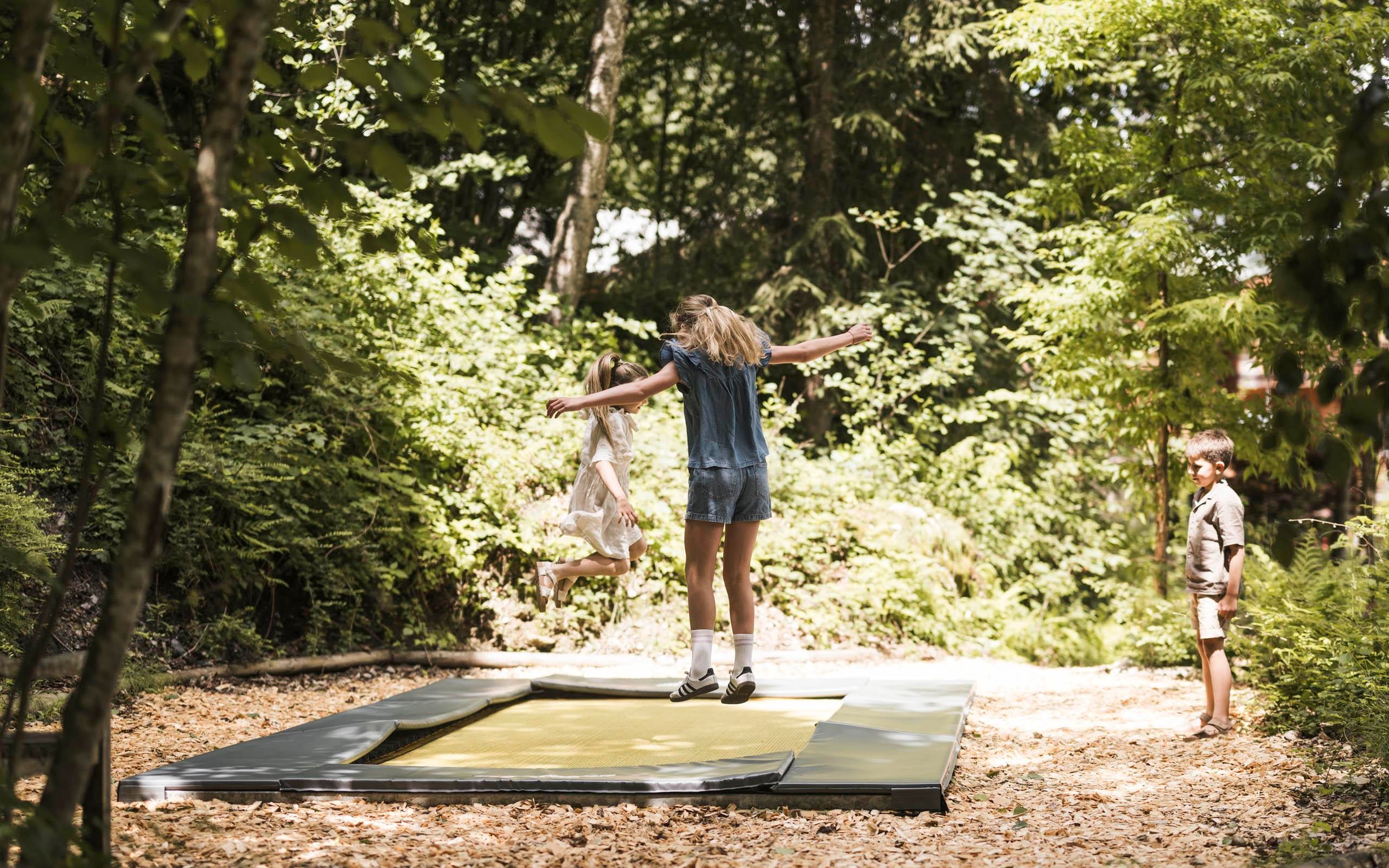 Ein Mädchen springt fröhlich auf einem Trampolin, während ein Junge daneben zusieht. Die Umgebung ist von Bäumen und Laub umgeben.