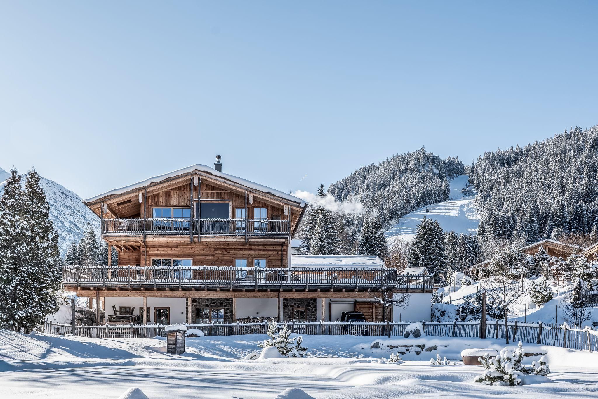 Ein charmantes Chalet im Winter mit Schnee bedeckt. Die umliegenden Berge sind mit Tannenbäumen versehen und der Himmel ist klar.