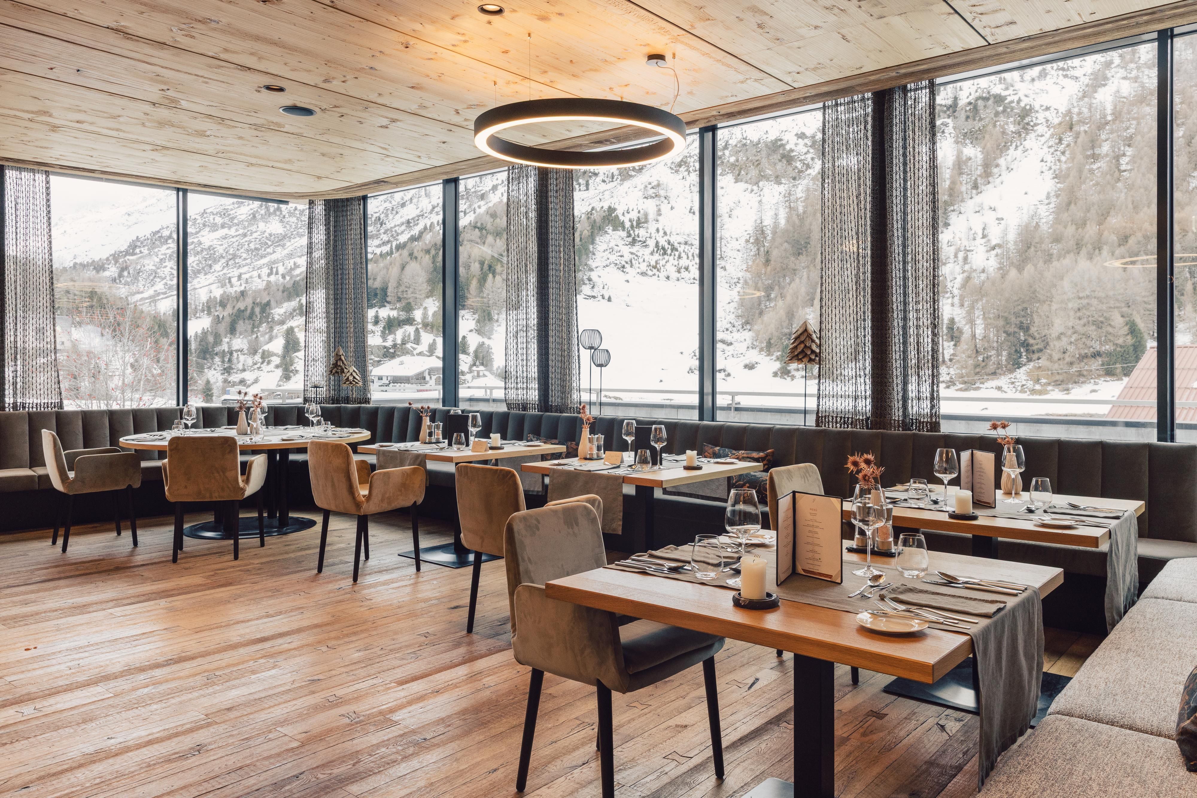 Ein modernes Restaurant mit großen Fenstern und Blick auf schneebedeckte Berge. Die Einrichtung ist stilvoll mit eleganten Tischen und Stühlen.