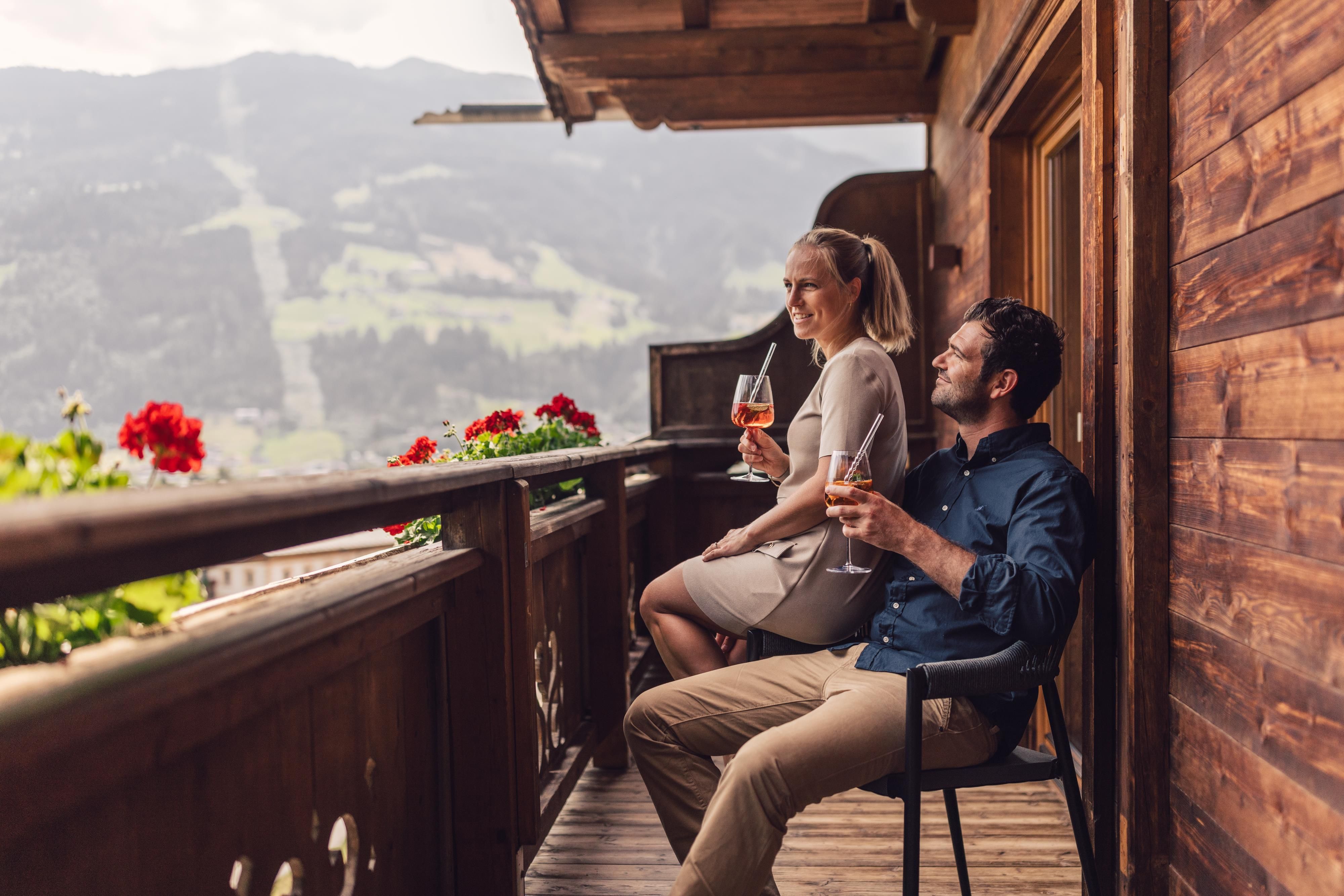 Ein Paar sitzt entspannt auf einem Balkon und genießt Getränke. Im Hintergrund sind die Berge und die Landschaft zu sehen.