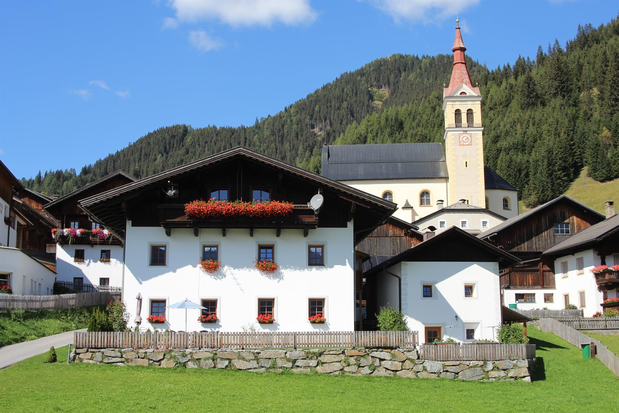 Sommeransicht vom Kleinbacherhof mit Bauernhaus und Kornkasten, vorne ist eine grüne Wiese, eine Steinmauer mit Holzzaun umfriedet den Garten, der dunkle Holzbalkon ist mit roten Blumen geschmückt, um den Kleinbacherhof herum sind Nachbarhäuser zu sehen, im Hintergrund eine Kirche.
