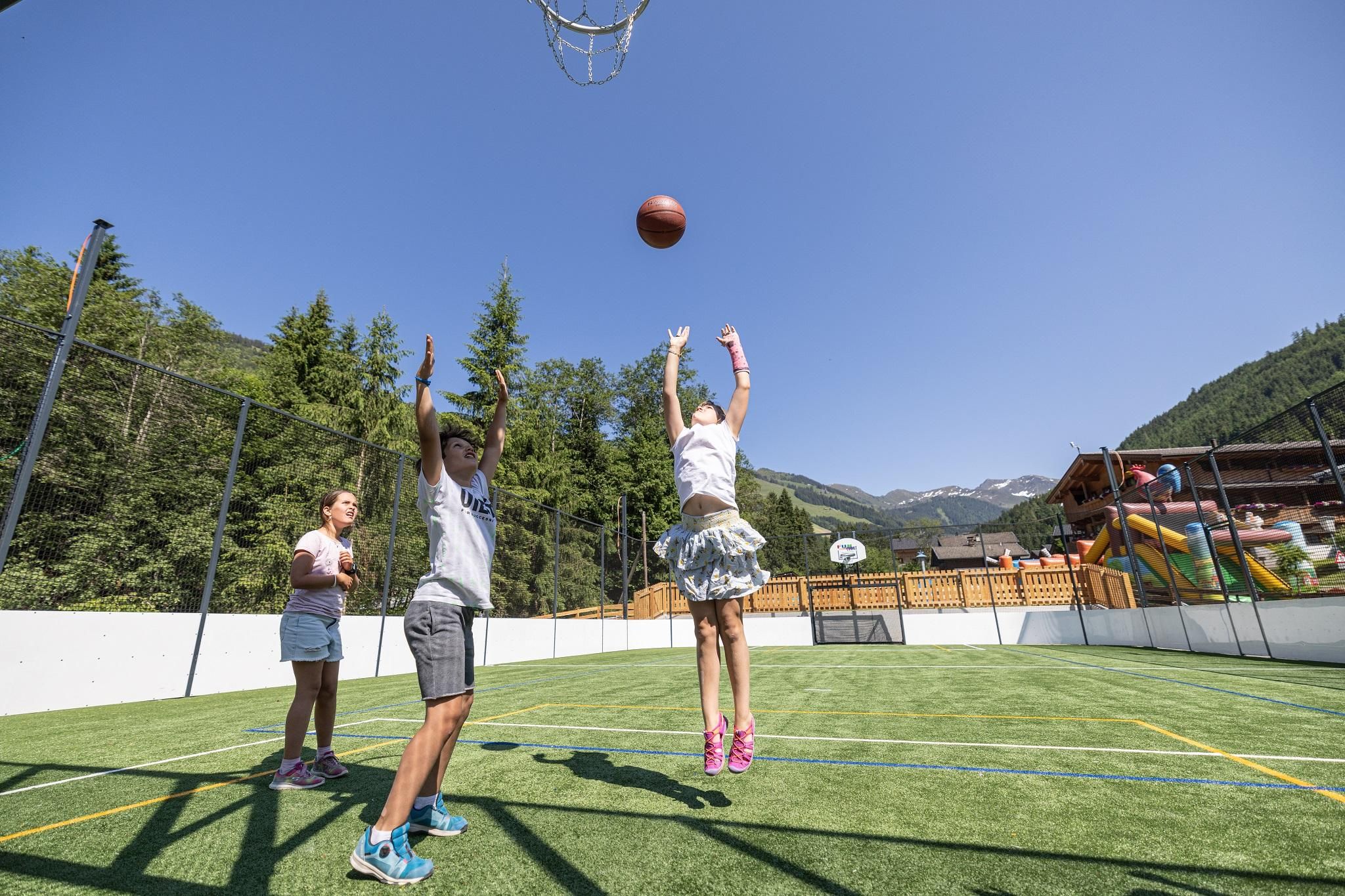 Ein Basketballplatz im Freien mit Kindern, die spielen. Im Hintergrund sind Bäume und ein Freizeitbereich sichtbar.