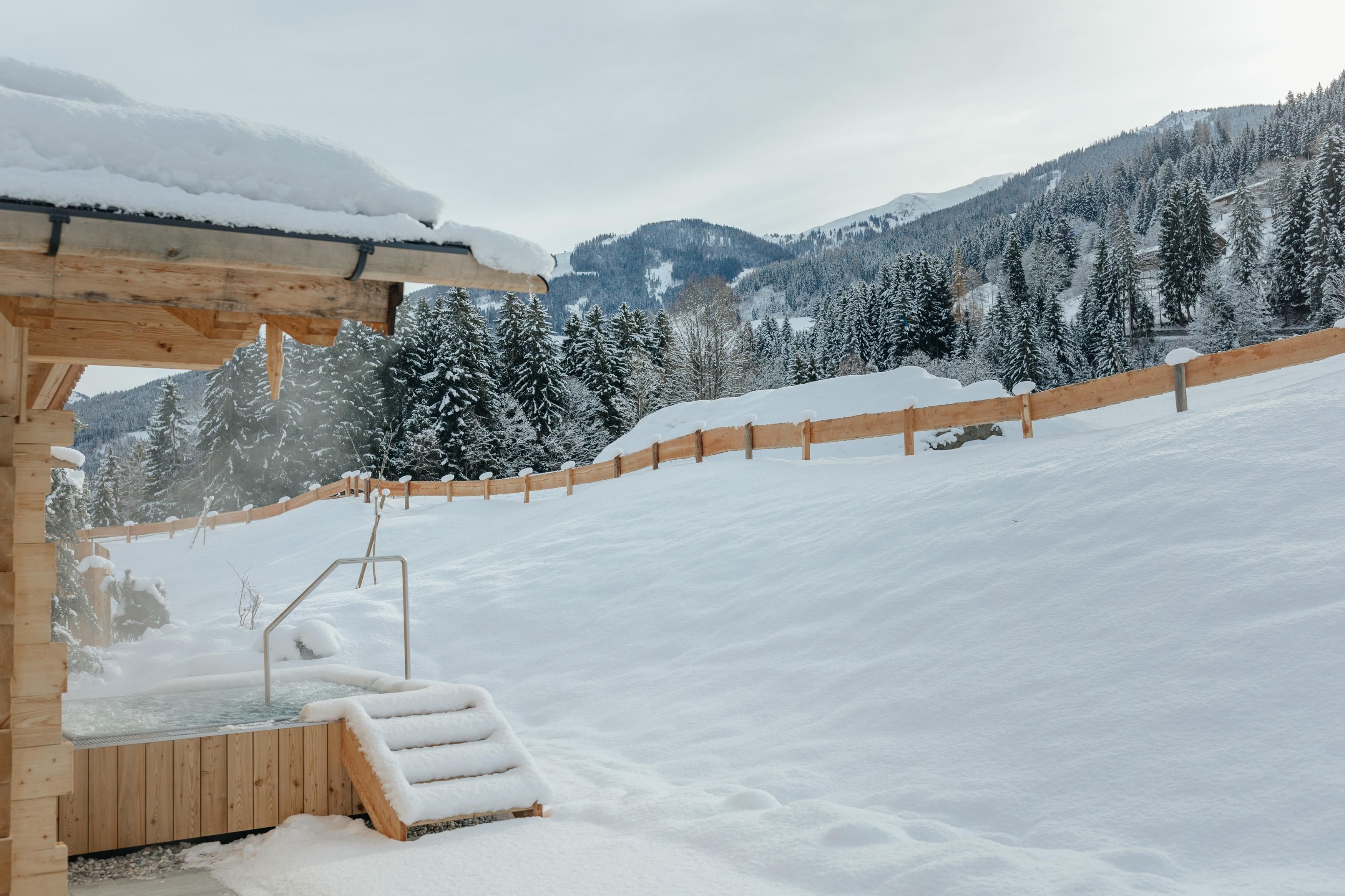 Ein verschneiter Landschaft mit einem Holzgebäude und einer Treppe. Im Hintergrund sind schöne Berge und Tannenbäume zu sehen.
