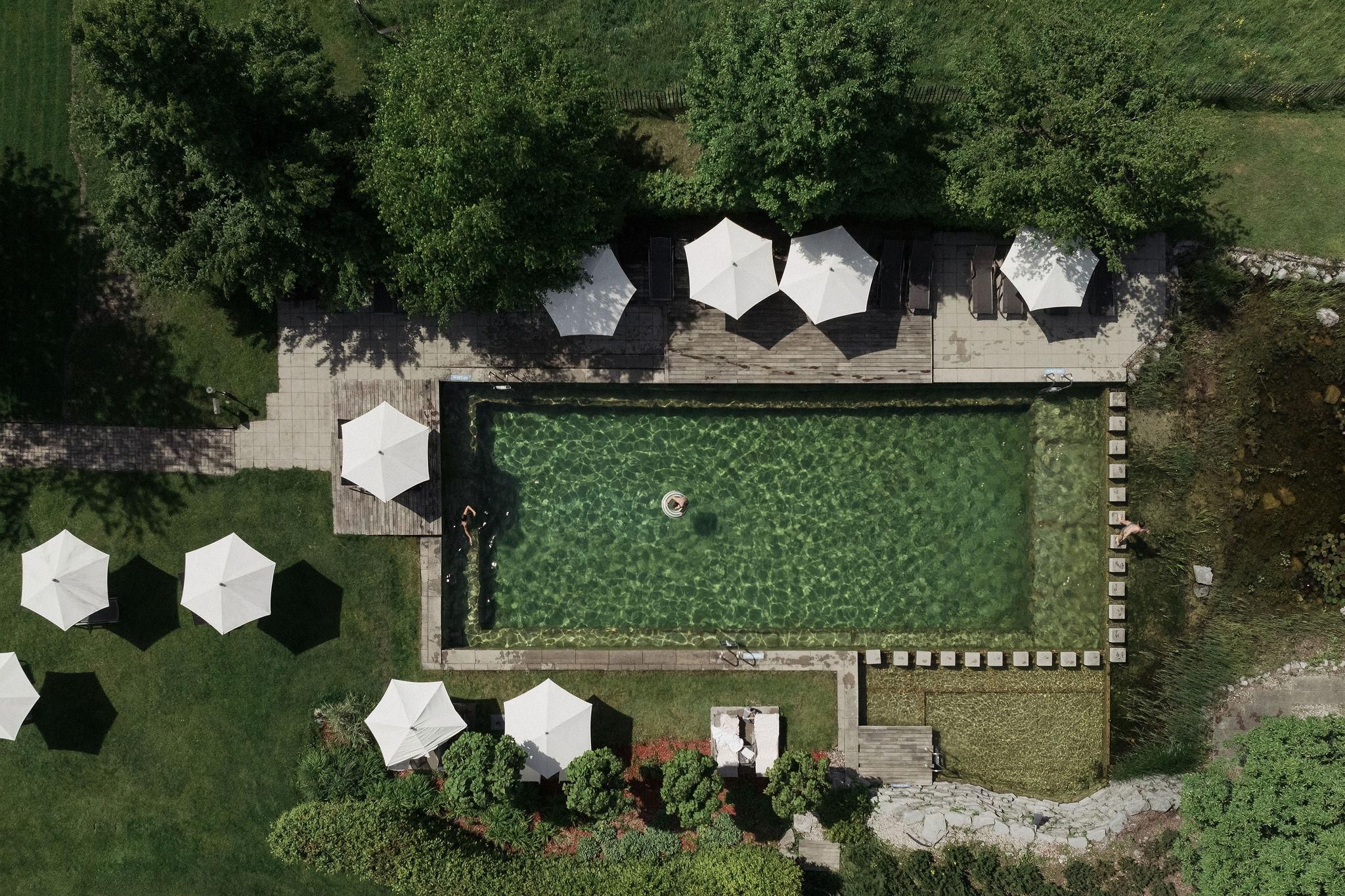 Ein schöner Pool umgeben von Bäumen und Liegestühlen. Weiße Sonnenschirme stehen auf der Terrasse.