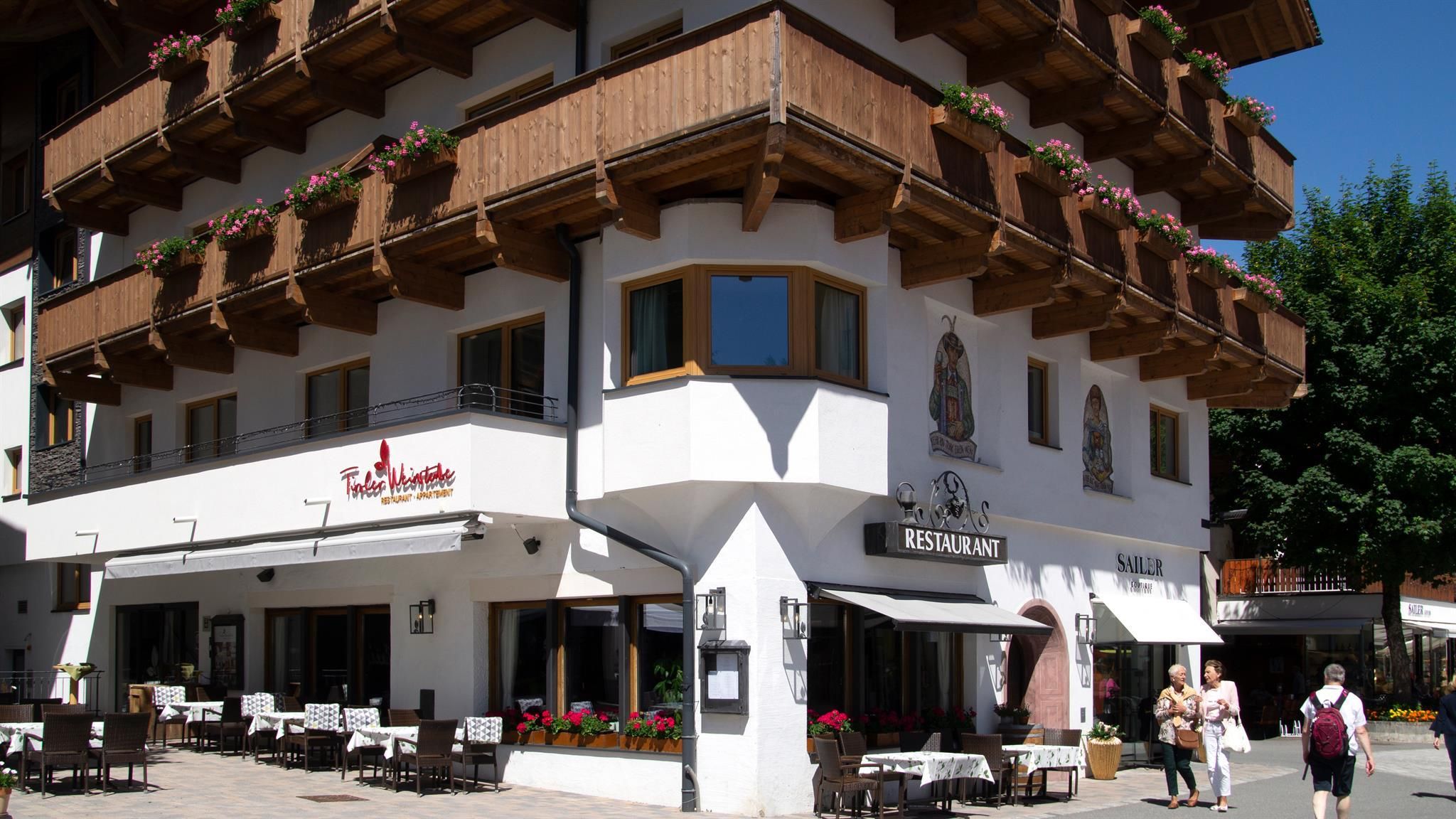 Ein schönes Restaurantgebäude mit Holzverkleidung und Blumenkästen. Menschen gehen entspannt auf der Straße vorbei.