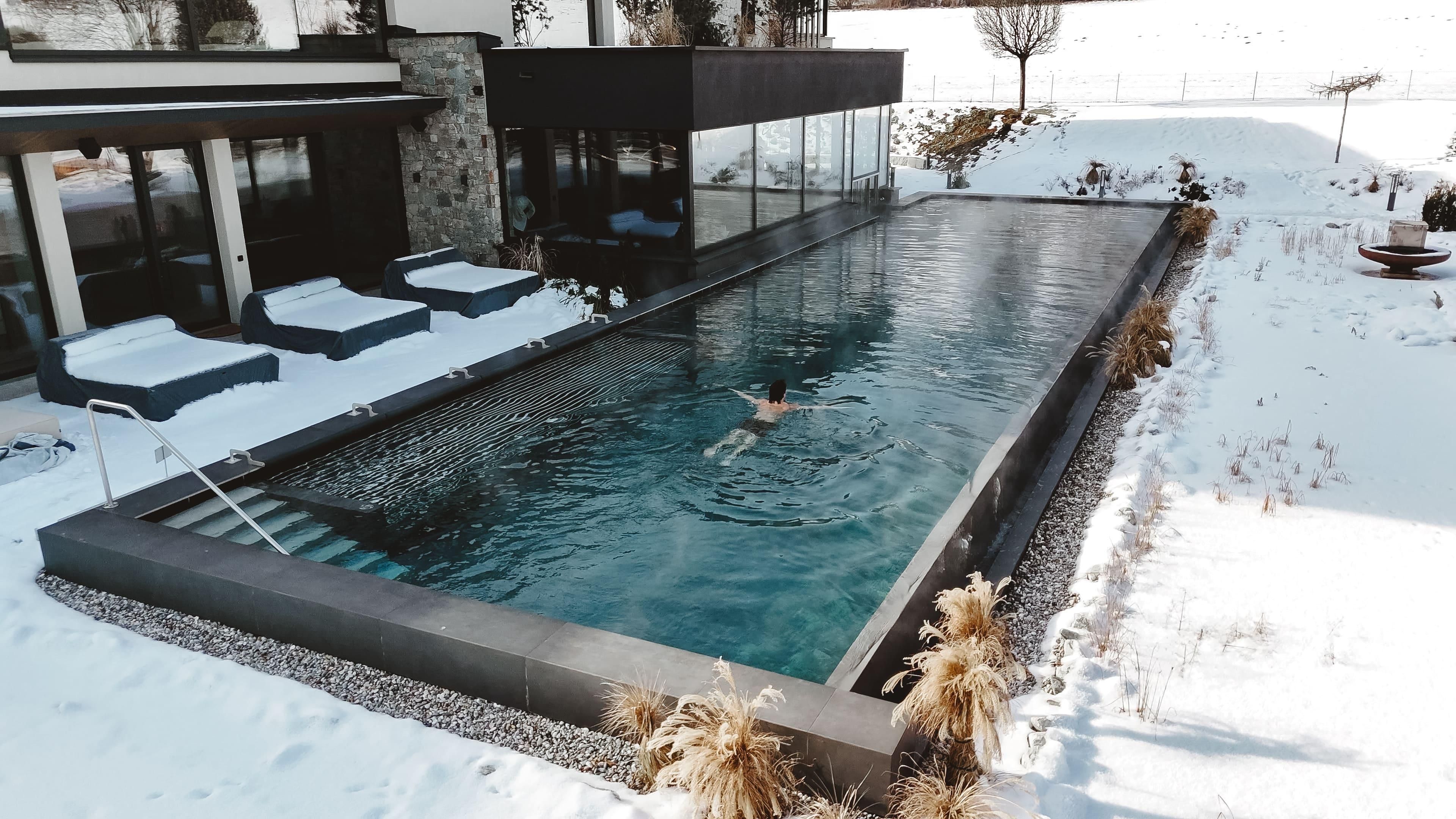 Ein moderner Pool, umgeben von Schnee, ideal zum Entspannen. Eine Person schwimmt im klaren Wasser, während ruhige Winterlandschaft im Hintergrund zu sehen ist.