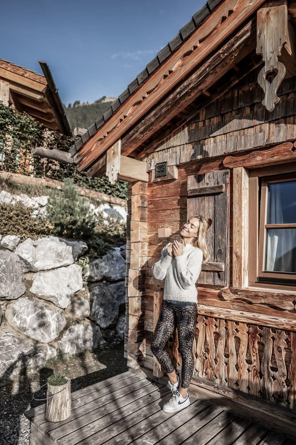 Eine junge Frau steht entspannt vor einem Chalet. Im Hintergrund sind Felsen und grüne Landschaft zu sehen.