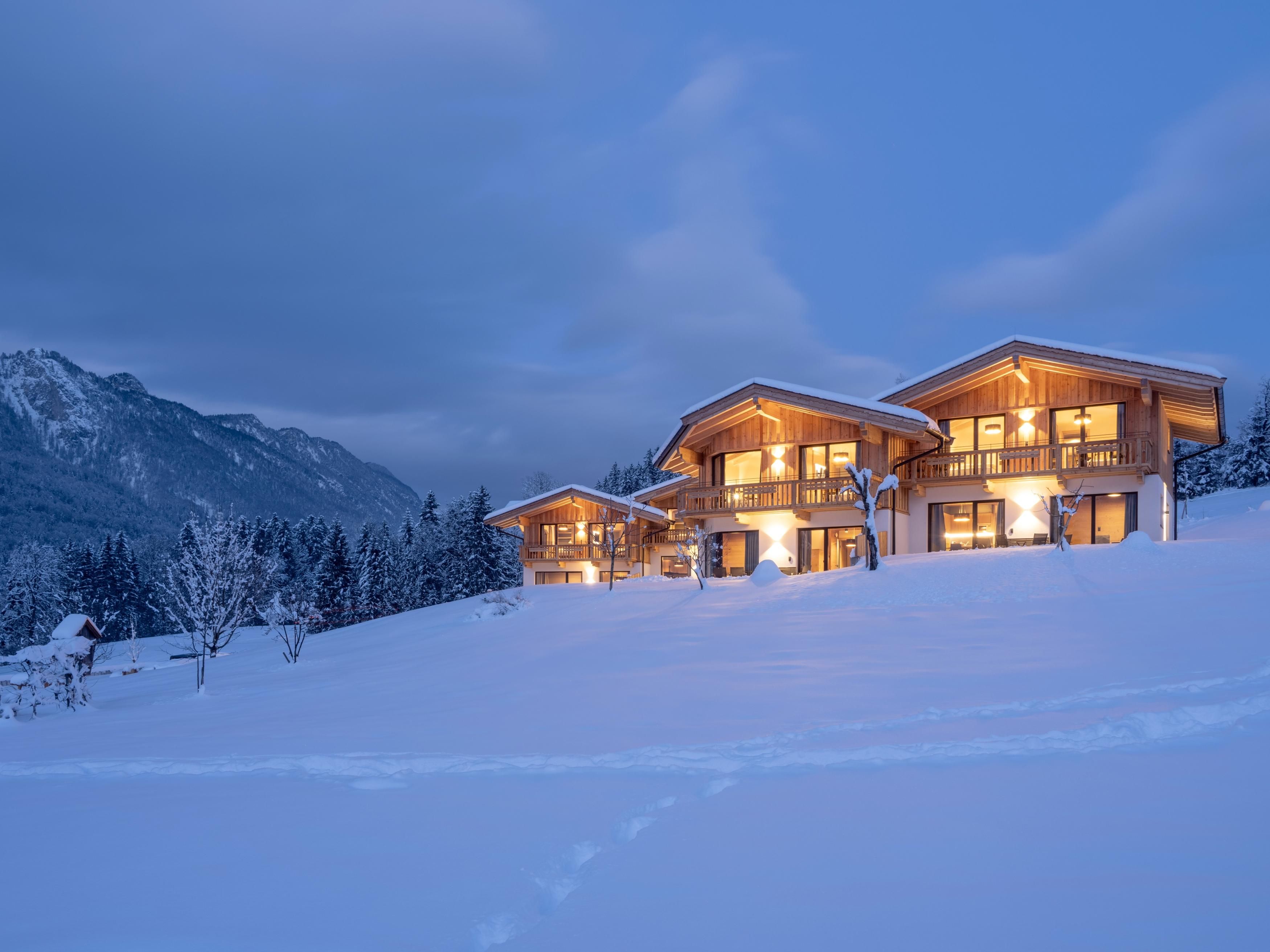 Eine gemütliche Chalet-Anlage im Schnee mit warmen Lichtern, umgeben von Bergen. Der Winterhimmel ist blau und bewölkt.