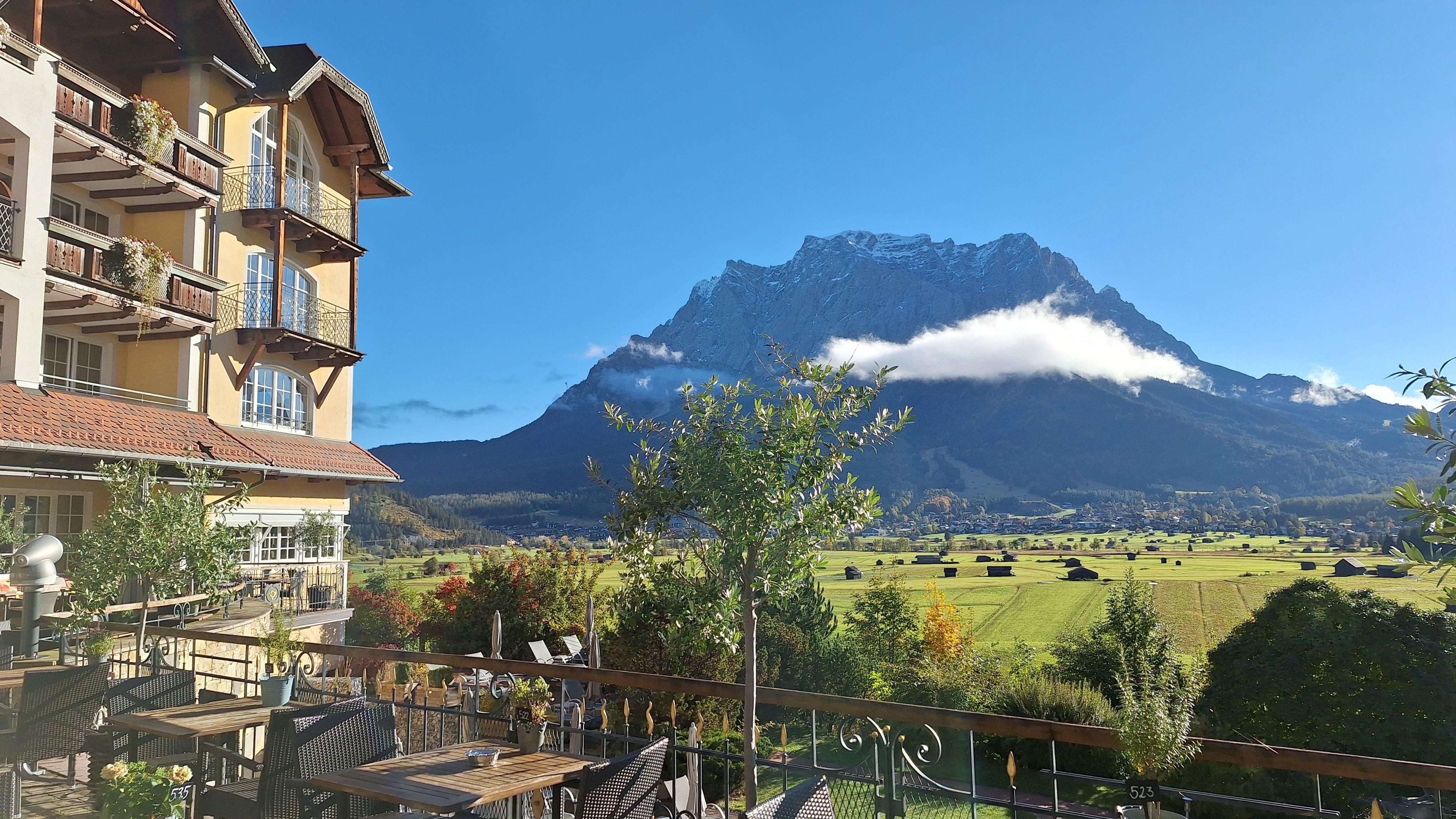 Eine malerische Landschaft mit einem beeindruckenden Berg im Hintergrund. Im Vordergrund sind einige Tische auf einer Terrasse und grüne Wiesen zu sehen.