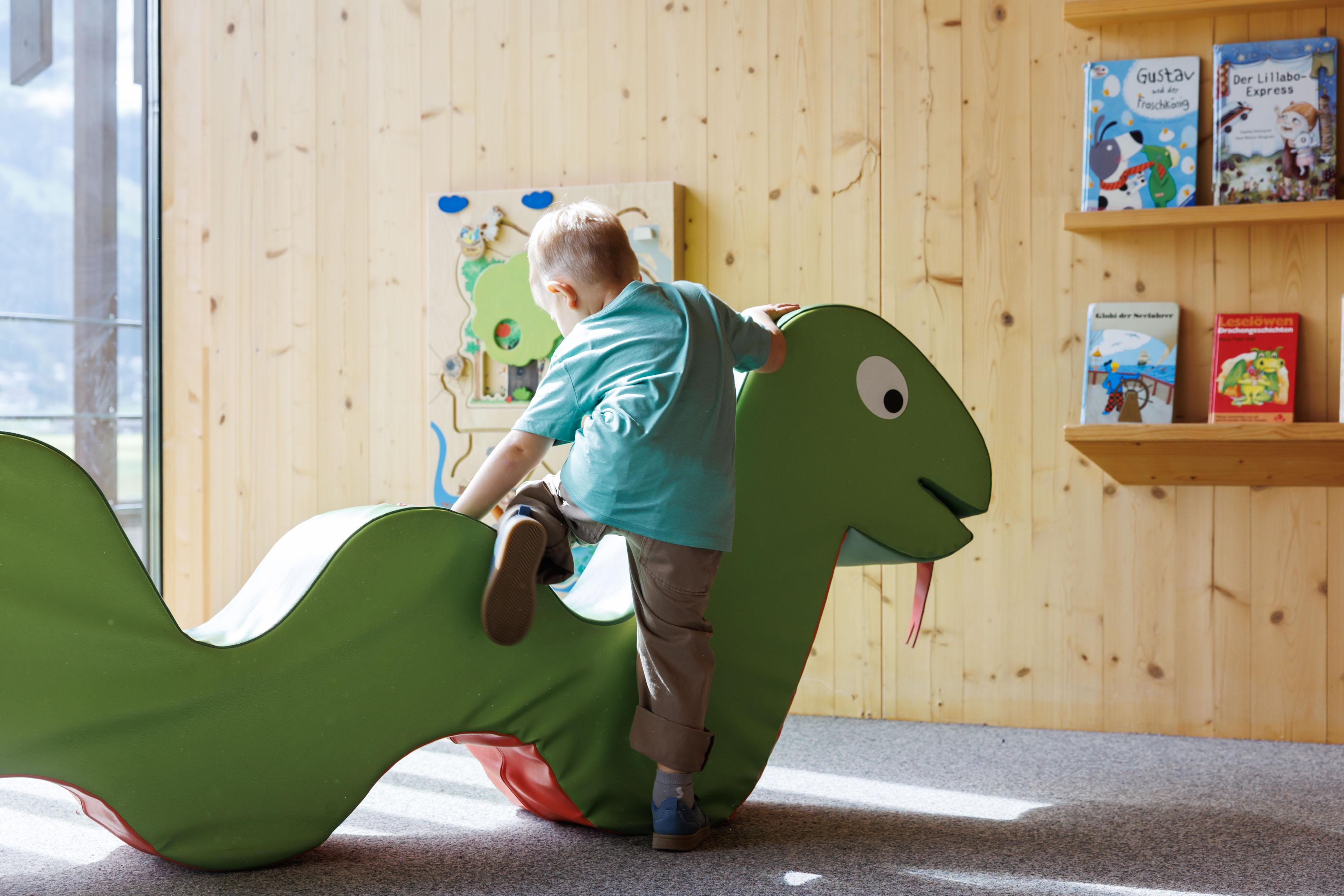 Ein Kind spielt auf einer grünen Schlangenfigur in einem hellen Raum. Im Hintergrund sind Regale mit Kinderbüchern zu sehen.