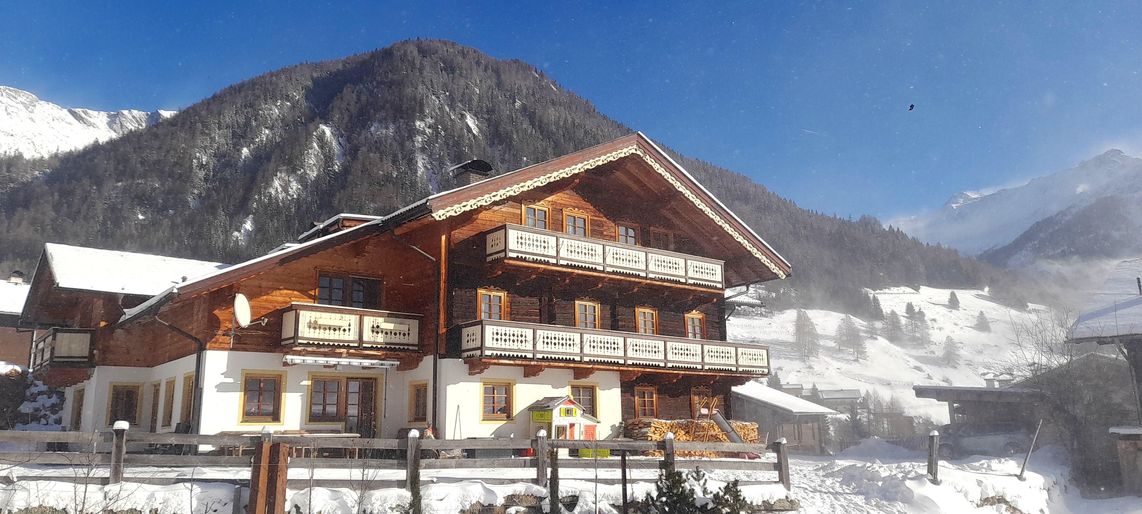 Ein charmantes Alpenhaus im Schnee mit mehreren Balkonen. Im Hintergrund ragen majestätische Berge unter klarem Himmel empor.