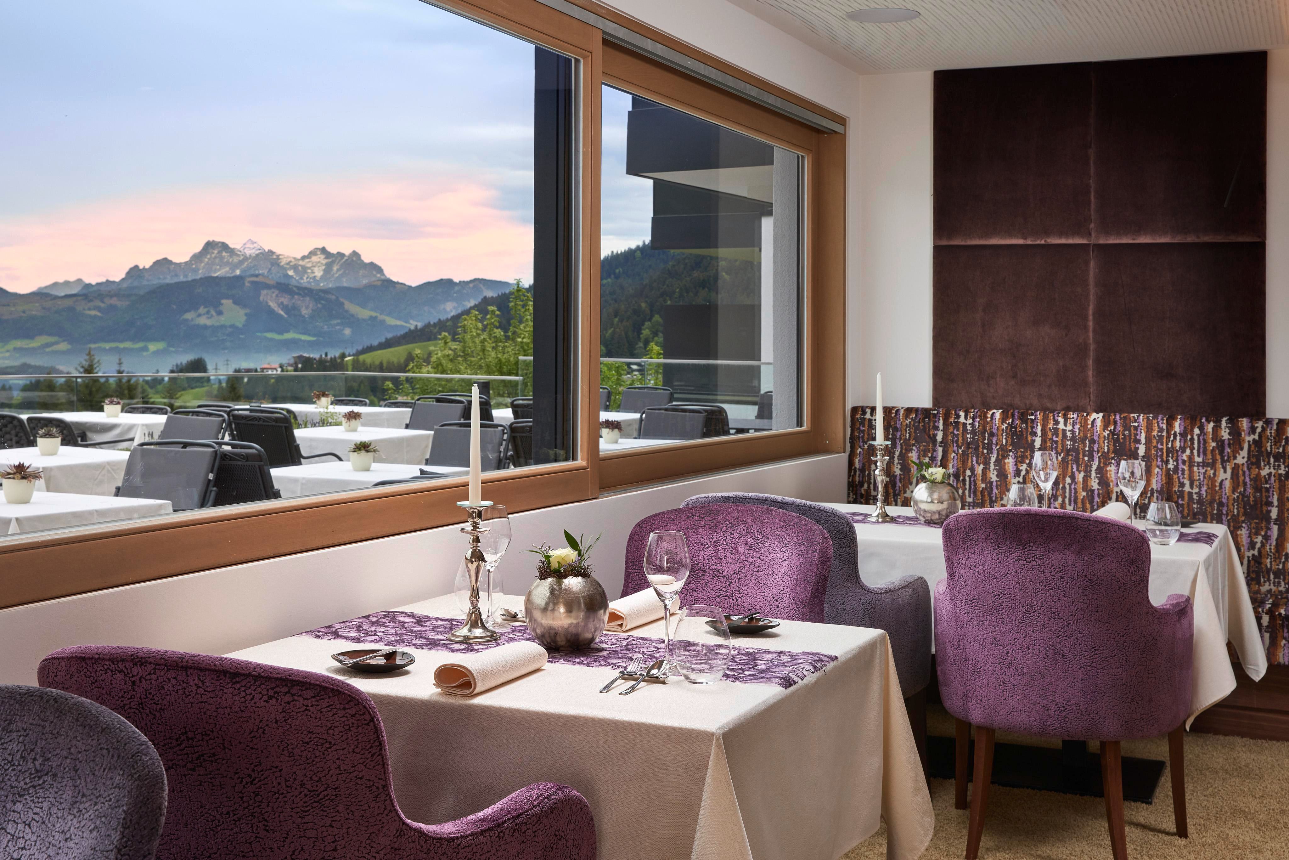 Ein elegantes Restaurant mit lila Stühlen und Tischdekoration. Im Hintergrund sind majestätische Berge und ein schöner Himmel sichtbar.