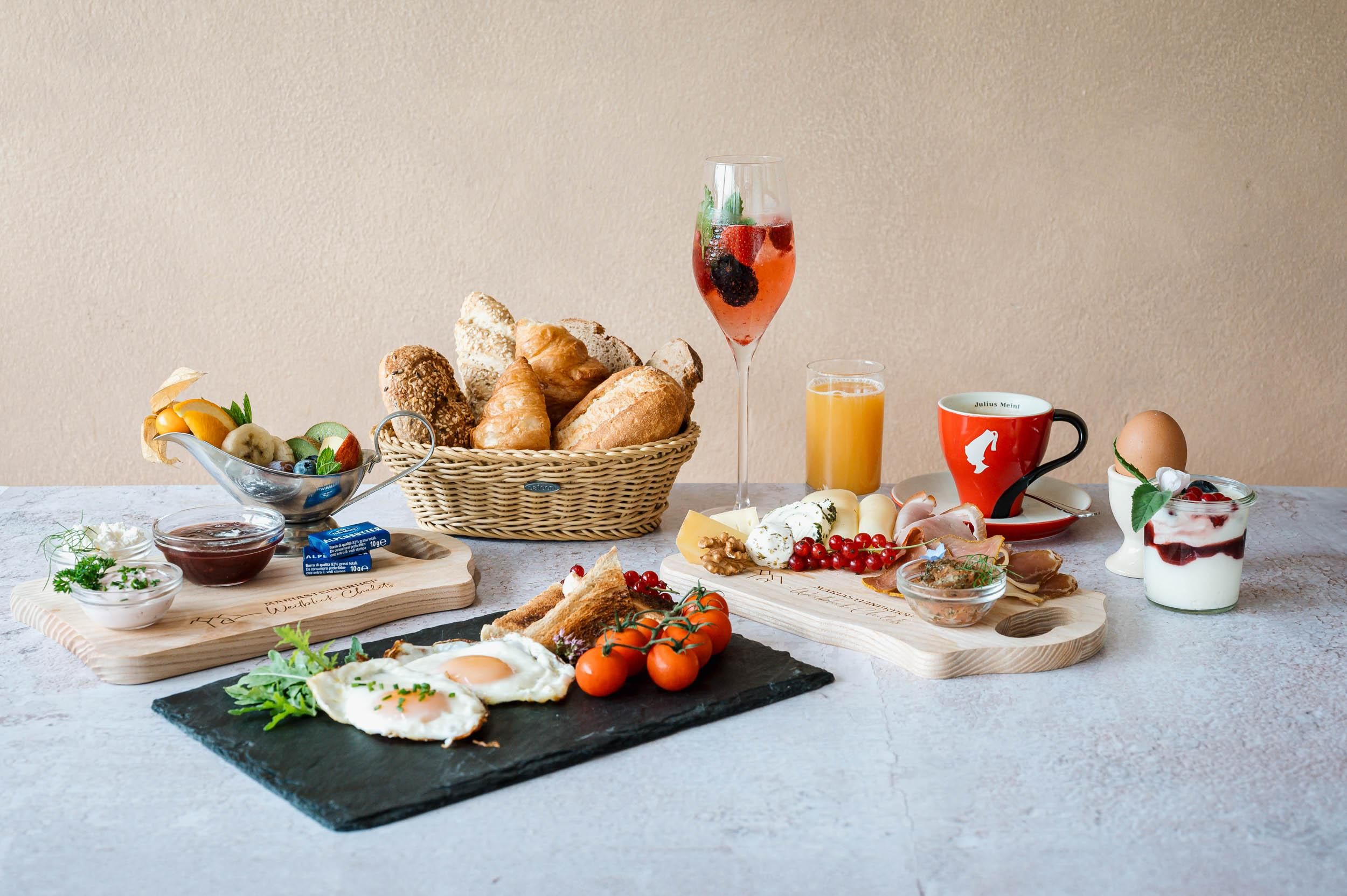 Ein reichhaltiges Frühstücksbuffet mit verschiedenen Brötchen, Eiern, Tomaten und einer Auswahl an Getränken. Dazu frisches Obst und süße Aufstriche.