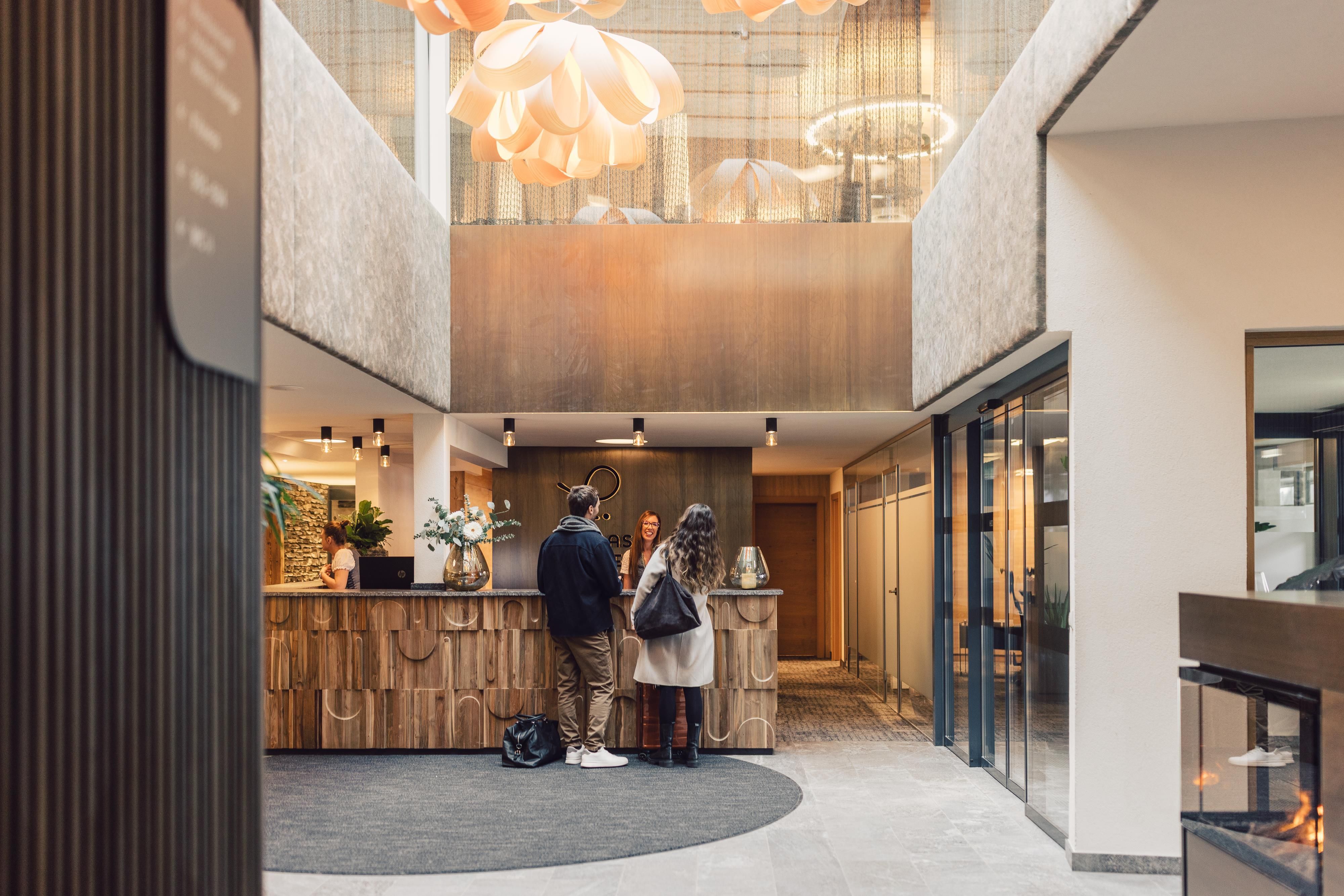 Eine moderne Hotel Lobby mit einem Empfangstresen aus Holz. Zwei Gäste stehen am Empfang, während eine Mitarbeiterin ihnen hilft.