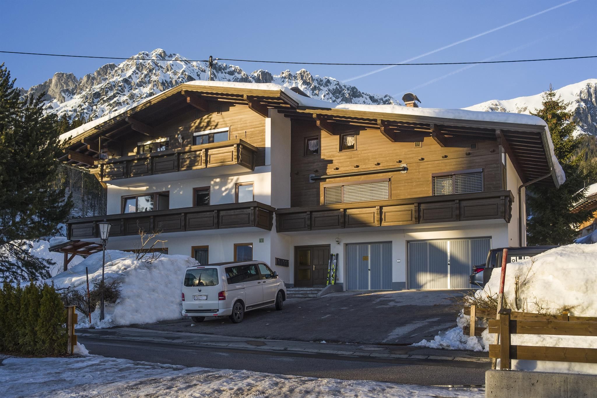 Ein modernes Holzhaus in einer schneebedeckten Landschaft mit Bergen im Hintergrund. Es gibt einen Parkplatz und klare, blaue Himmel.