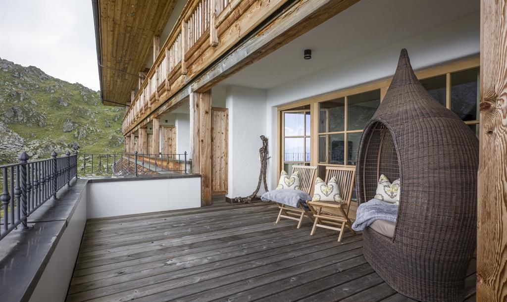 Eine gemütliche Veranda mit einem geflochtenen Sitzplatz und zwei Schaukelstühlen. Im Hintergrund sind grüne Hügel und ein Holzgebäude zu sehen.
