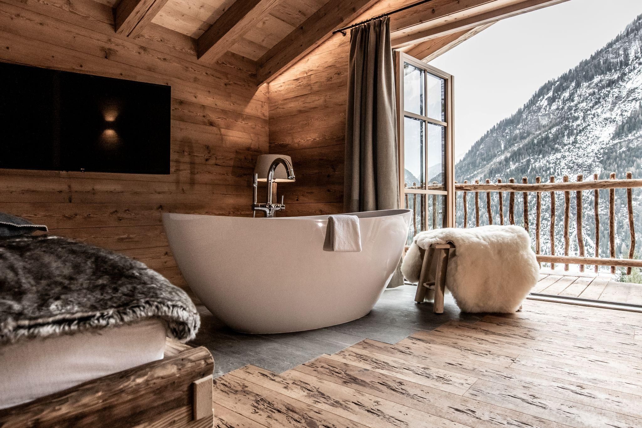 Ein modernes Badezimmer in einem Chalet mit einer freistehenden Badewanne. Die große Fensterfront bietet einen Blick auf schneebedeckte Berge.
