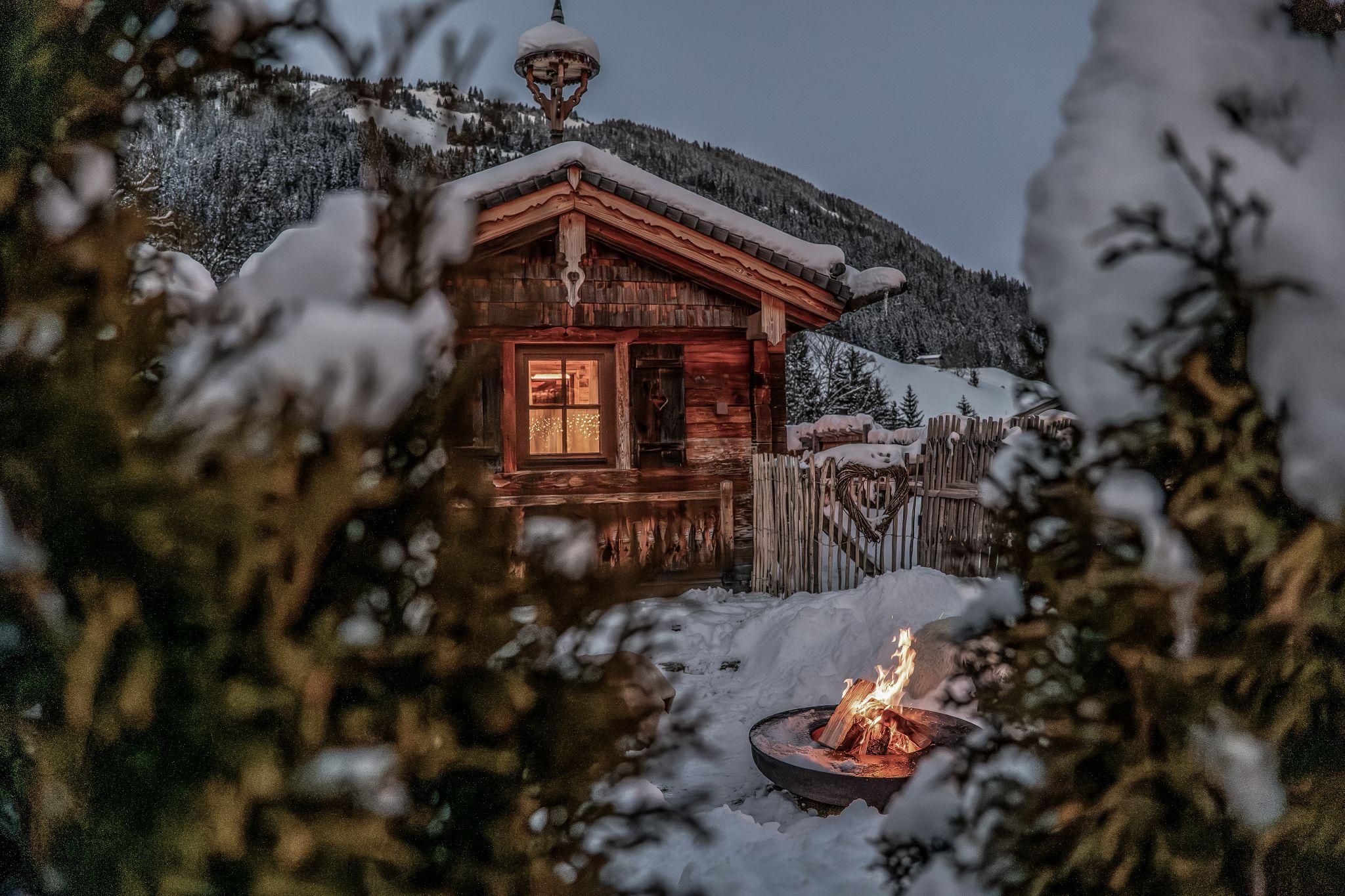 Eine gemütliche Chalet im Schnee mit einem warmen Feuer im Vordergrund. Die Umgebung ist winterlich und malerisch.