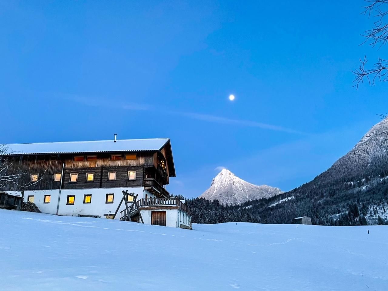 Eine malerische Berghütte im Schnee, umgeben von majestätischen Bergen. Der klare Himmel und der Mond verleihen der Szene eine ruhige Atmosphäre.