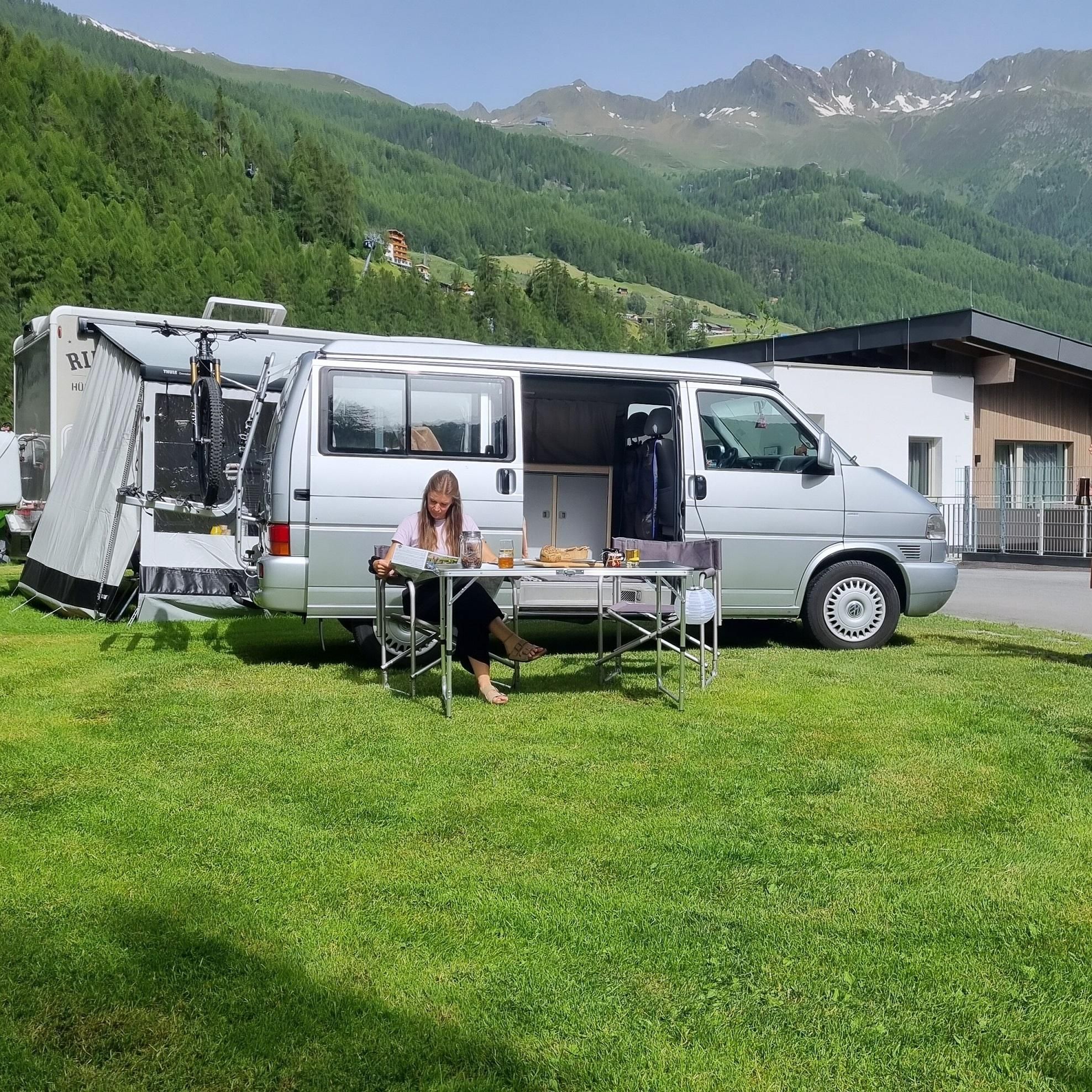 Ein Wohnmobil steht auf einer grünen Wiese mit Blick auf die Berge. Eine Frau sitzt an einem Tisch und genießt eine Mahlzeit im Freien.