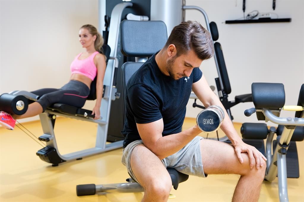Ein Mann hebt eine Hantel im Fitnessstudio, während eine Frau auf einem Trainingsgerät sitzt. Die Atmosphäre ist aktiv und sportlich.