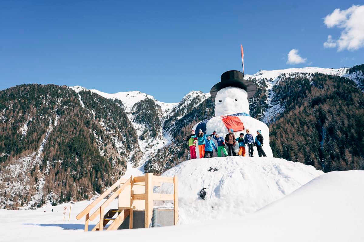Eine Gruppe von Kindern steht neben einem großen Schneemann in einer winterlichen Landschaft. Im Hintergrund sind schneebedeckte Berge und ein blauer Himmel zu sehen.