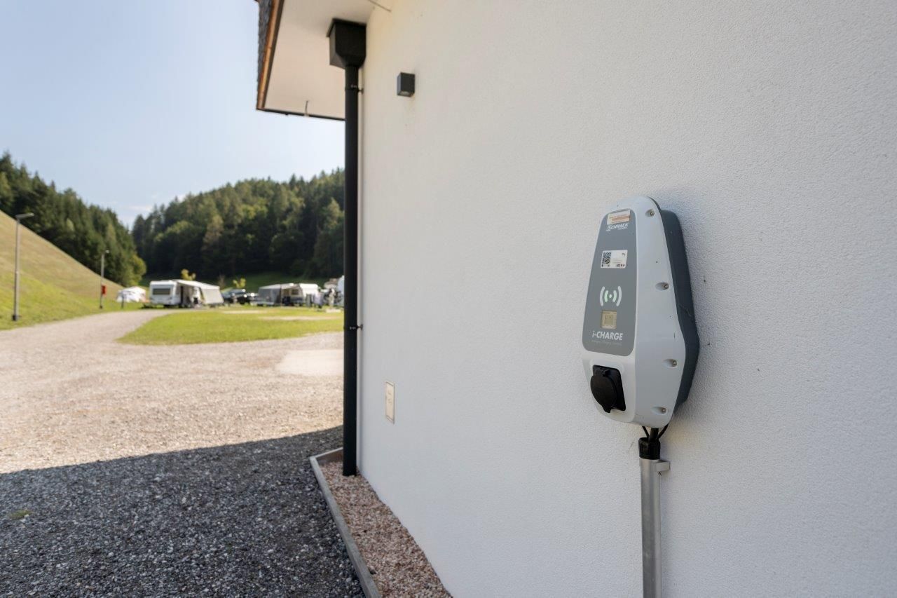 Eine Ladestation für Elektrofahrzeuge an einer Außenwand. Im Hintergrund sind Bäume und ein Weg sichtbar.