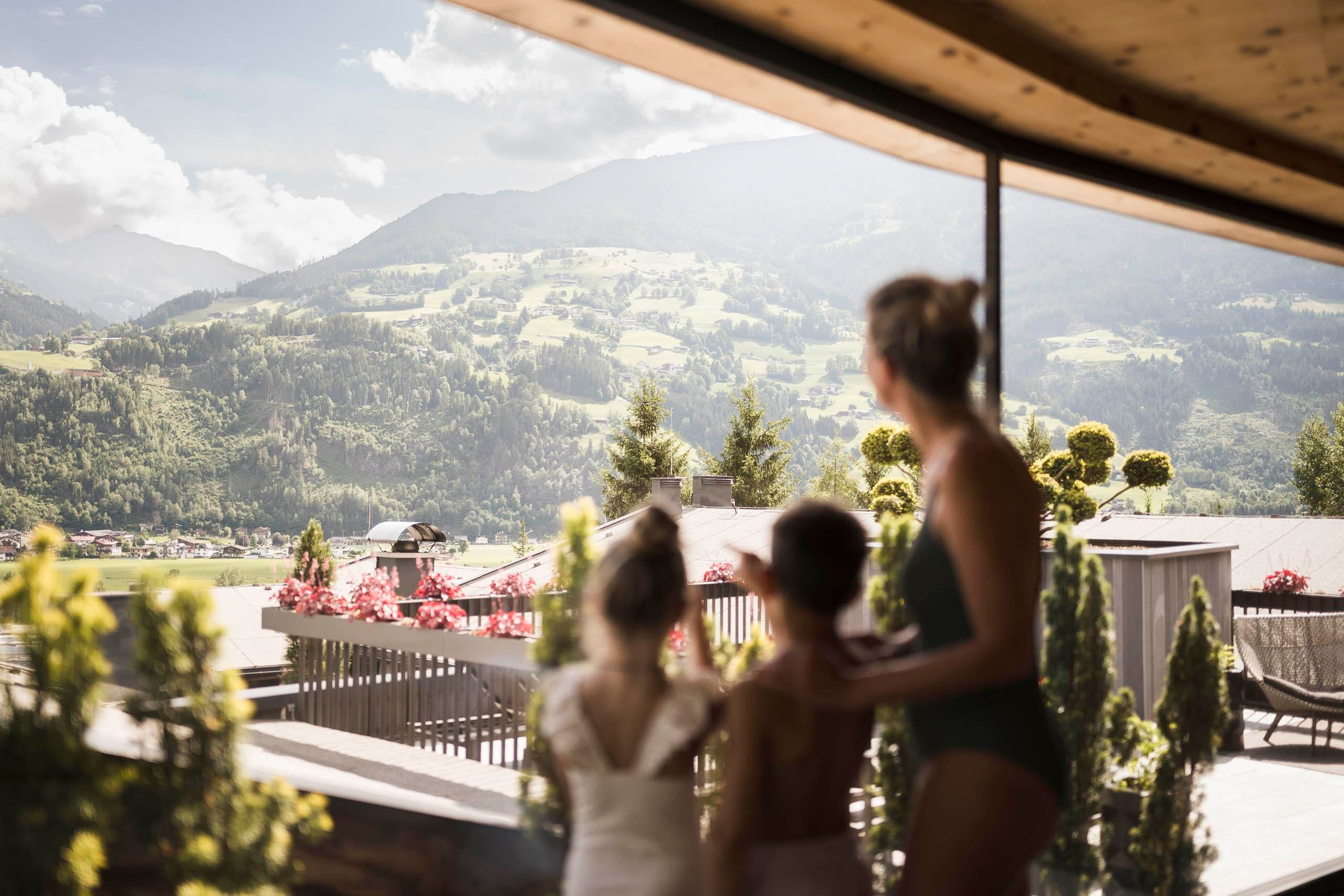 Eine Familie steht auf einer Terrasse und blickt auf eine wunderschöne Berglandschaft. Die Umgebung ist mit bunten Blumen und grünen Pflanzen geschmückt.