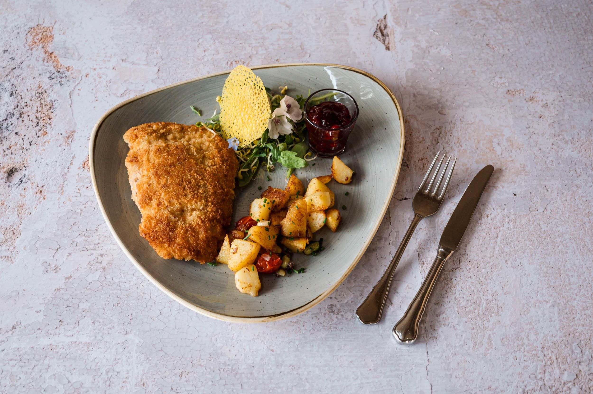 Ein gut angerichtetes Gericht mit paniertem Fisch, Beilagen aus Kartoffeln und Gemüse sowie einer kleinen Sauce. Das Essen ist auf einem eleganten Teller serviert.