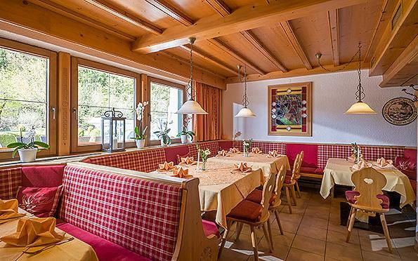 Ein gemütliches Restaurant mit Holzbalkendecke und roten Karostühlen. Die Tische sind mit gelben Tischdecken dekoriert und die Fenster bieten einen Blick ins Grüne.