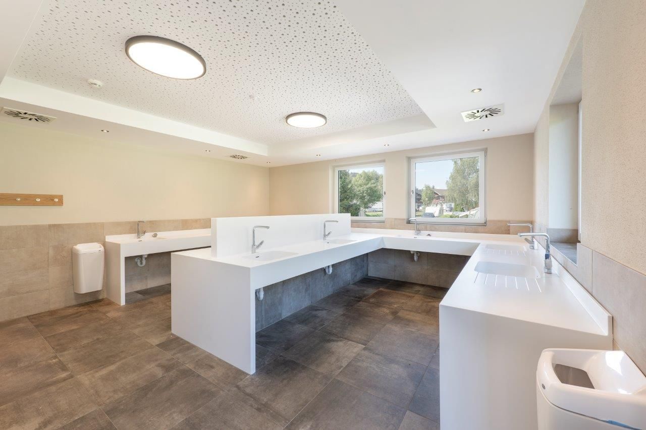 Ein modernes Badezimmer mit weißen Waschbecken und großzügigen Spiegeln. Große Fenster sorgen für viel Tageslicht.