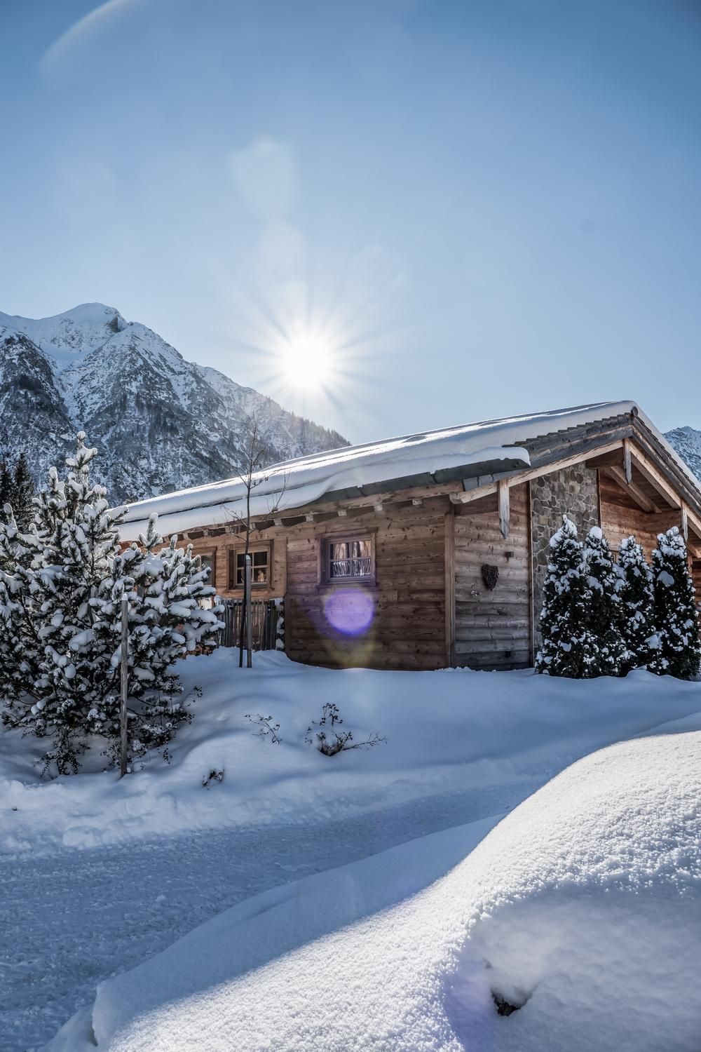 Eine gemütliches Chalet im Schnee, umgeben von schneebedeckten Bäumen. Die Sonne scheint hinter den Bergen hervor.