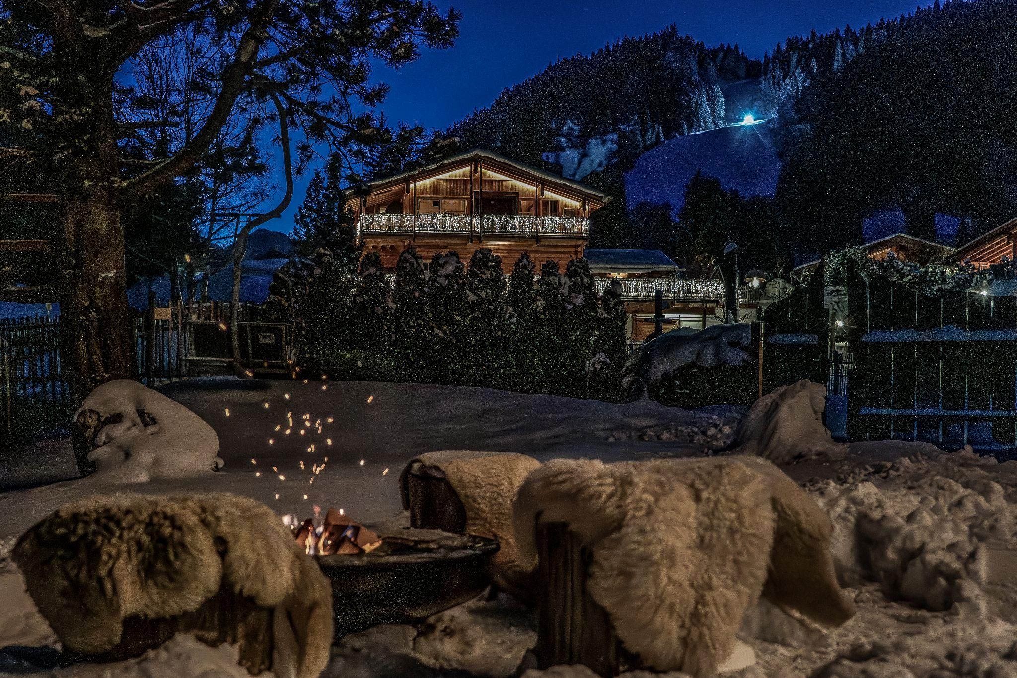 Ein gemütlicher Garten im Schnee mit einem Feuerplatz. Im Hintergrund ist ein wunderschönes Chalet und eine winterliche Berglandschaft zu sehen.