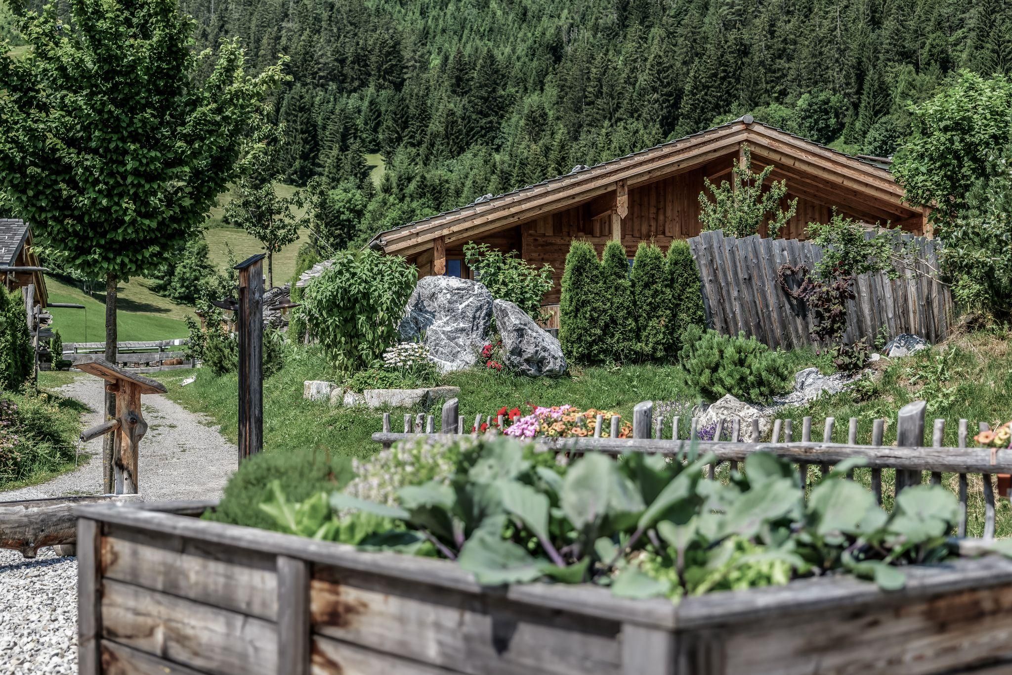 Ein gemütliches Chalet umgeben von grüner Landschaft und Bäumen. Im Vordergrund sind Blumen und ein Gemüsebeet zu sehen.