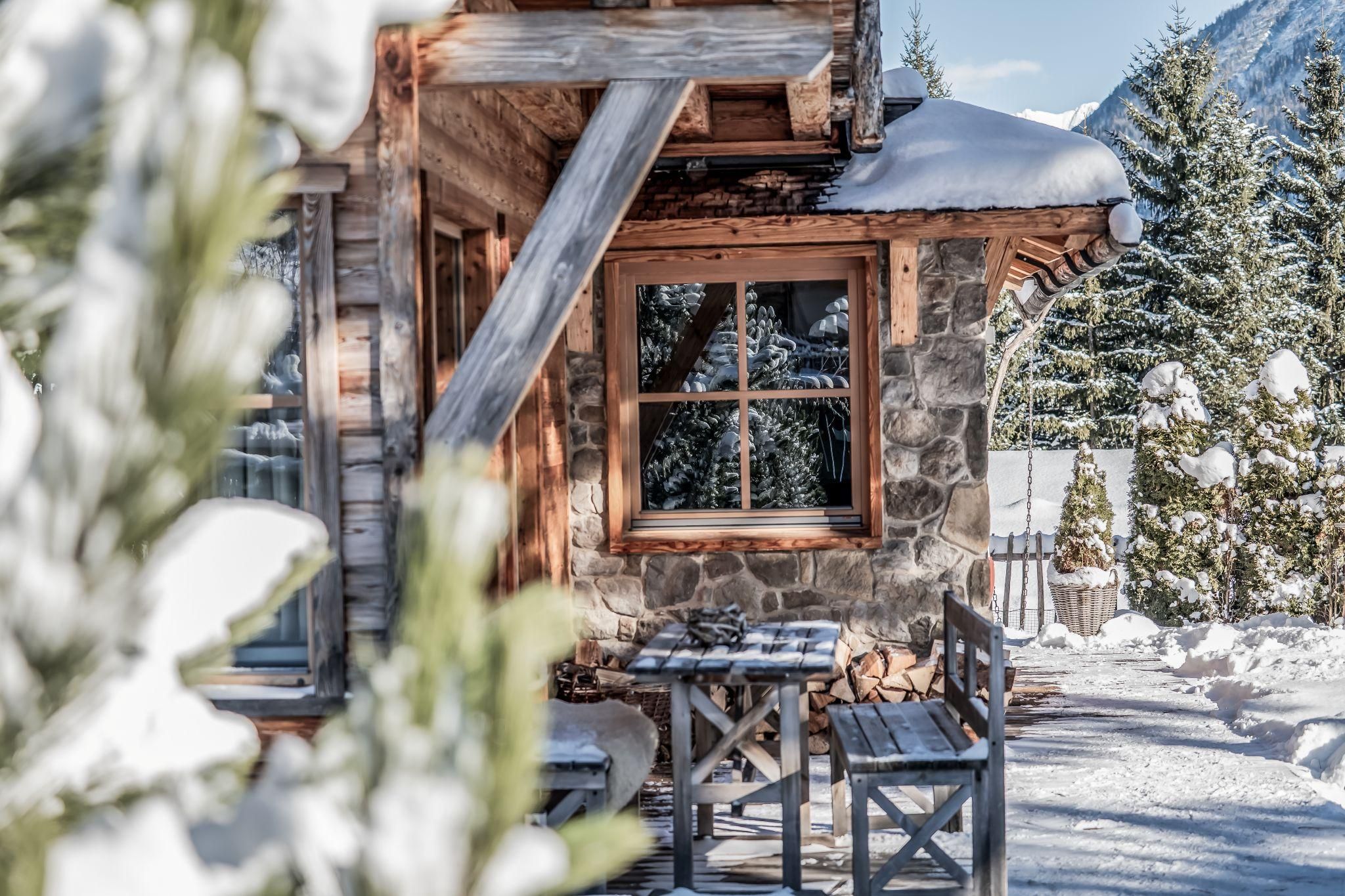 Ein gemütliches Chalet im Schnee mit einem großen Fenster. Umgeben von Tannenbäumen und einer winterlichen Landschaft.