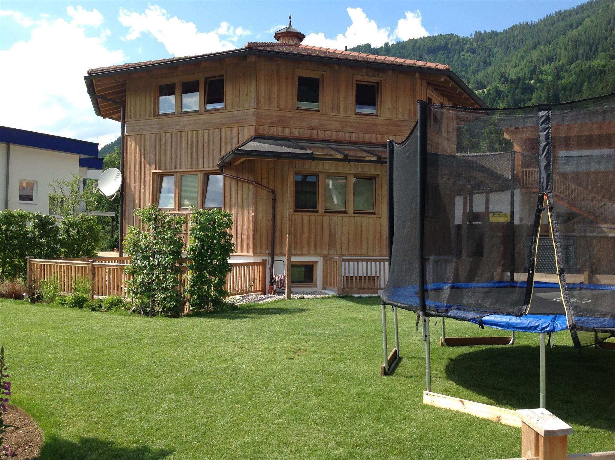 Ein gemütliches Holzhaus mit einem gepflegten Garten und einer Schaukel. Im Hintergrund sind grüne Berge zu sehen.