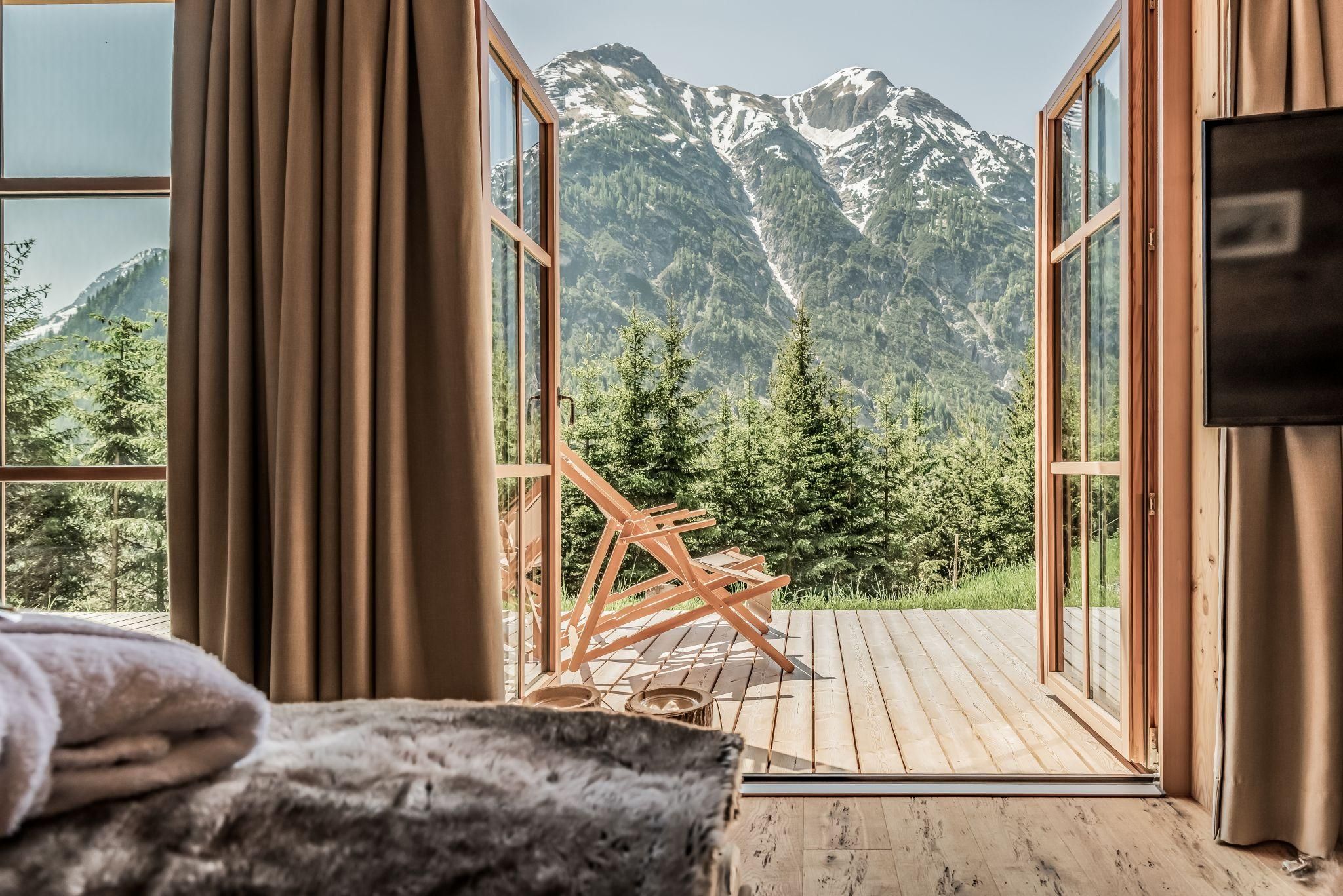 Ein gemütlicher Raum mit offener Tür, die zu einer Terrasse führt. Im Hintergrund sind majestätische Berge und grüne Bäume zu sehen.