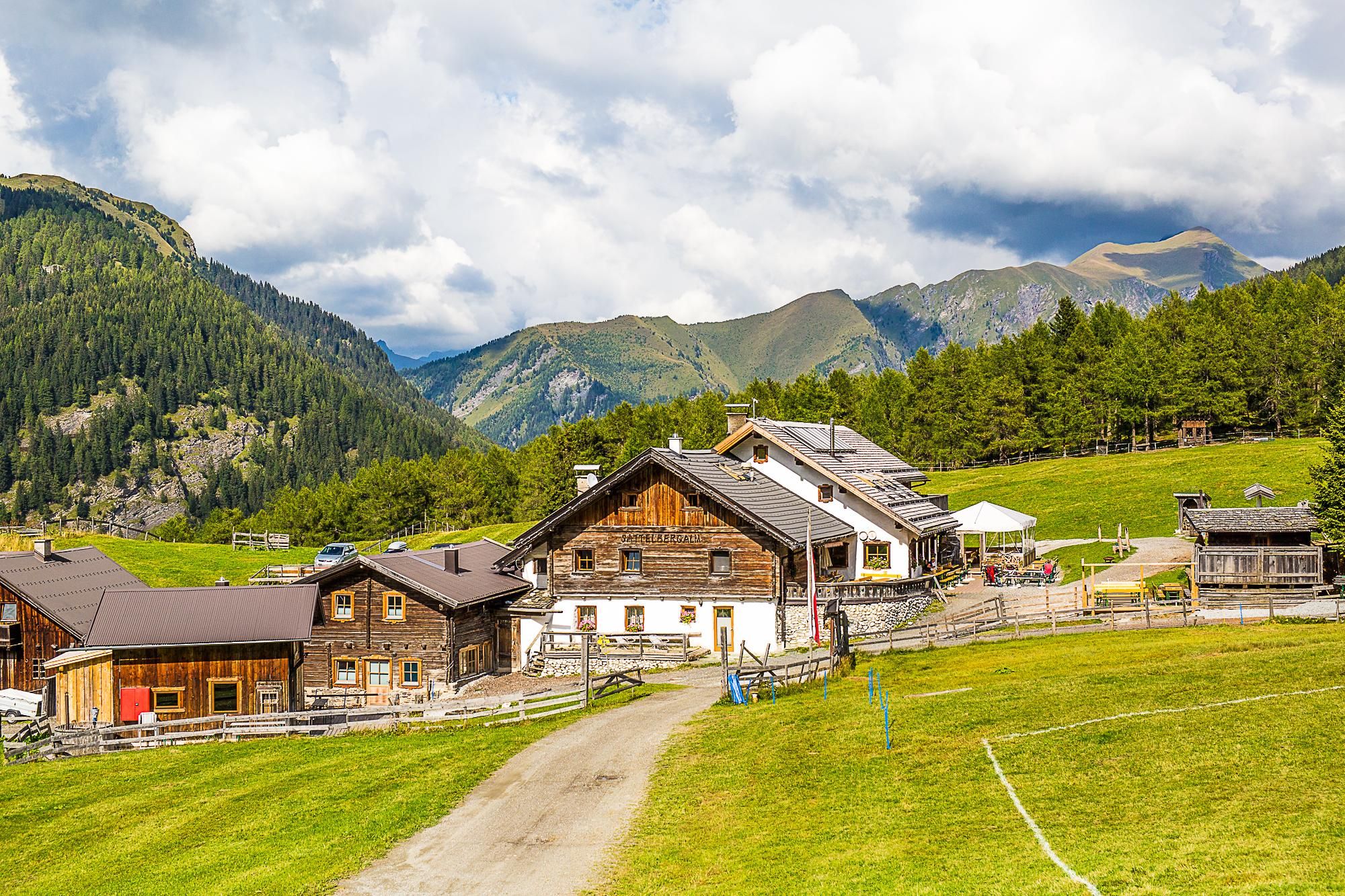Ein malerisches alpenländisches Dorf mit traditionellen Holzhäusern. Die Umgebung ist grün, umgeben von Bergen und einem klaren Himmel.