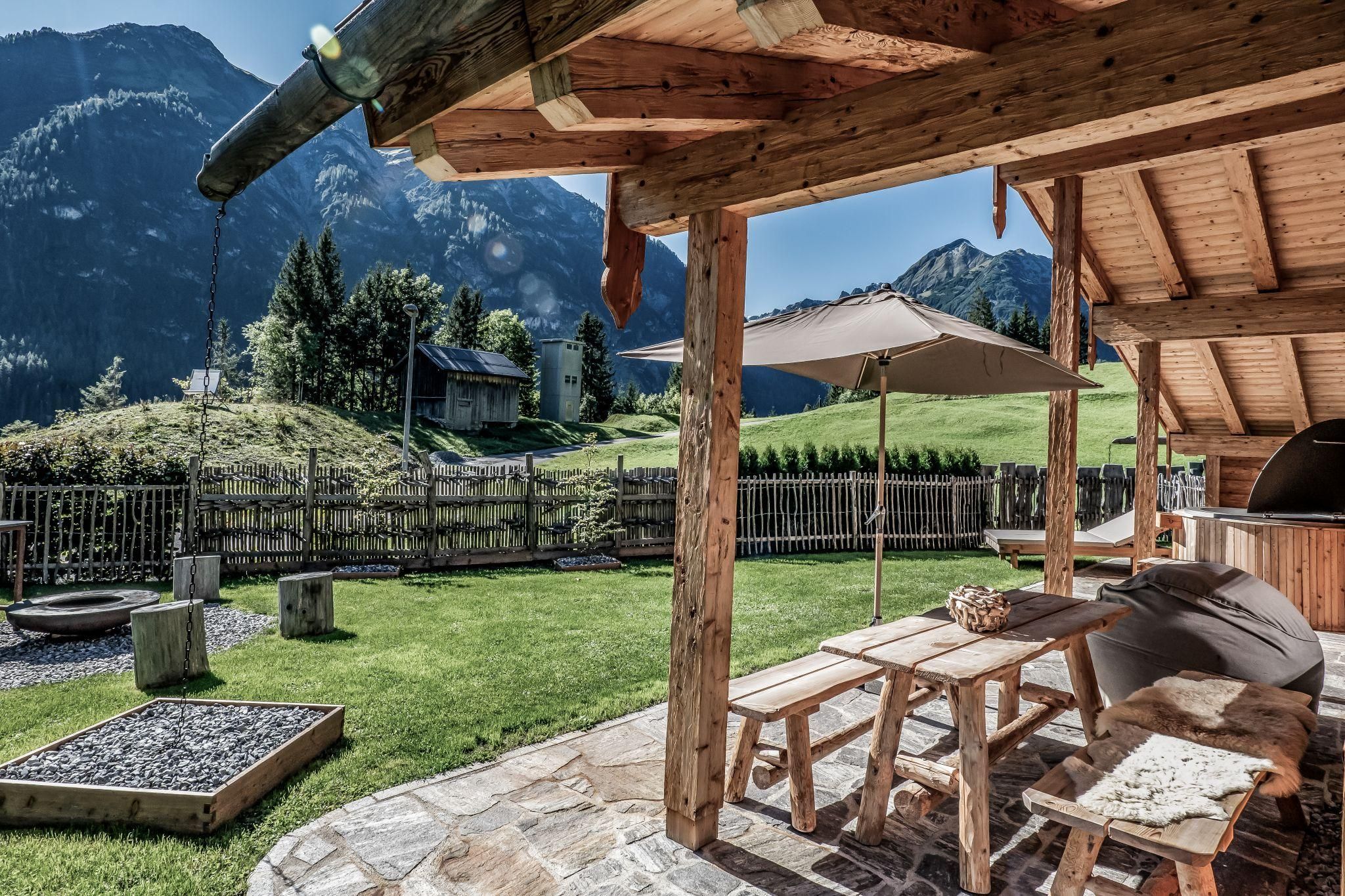 Ein gemütlicher Außenbereich mit einem Holztisch und Bänken. Im Hintergrund sind grüne Wiesen und Berge zu sehen.
